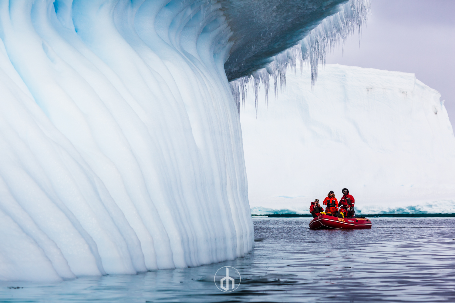 Three people in red survival suits and helmets on a small boat near a large iceberg with a curved top and hanging icicles, in icy waters under a cloudy sky.