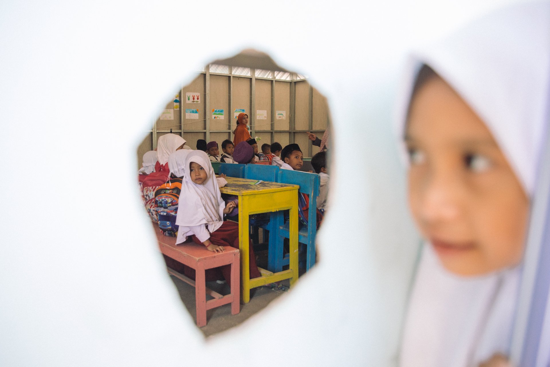 Children sitting on benches and at desks in a classroom, seen through a cutout in a white wall or object, with a woman standing at the back.