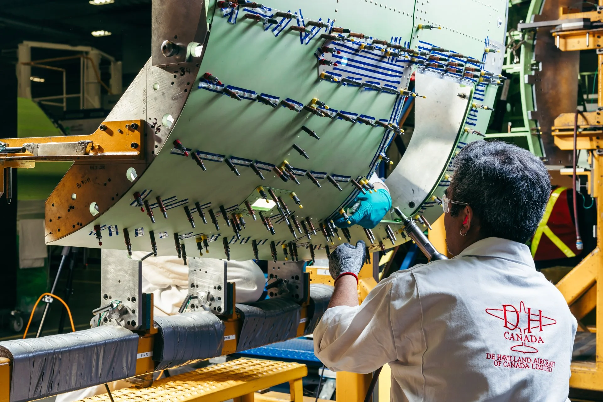 A technician working on an aircraft component at Dehavilland Aircraft of Canada Limited, wearing a white lab coat with the company's logo and safety glasses, in an industrial setting with machinery and tools around.