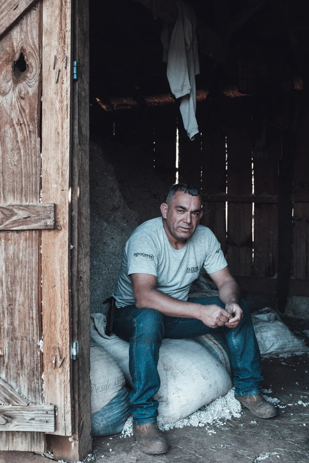 Man sitting on a sack inside a rustic wooden shed with sunlight filtering through gaps in the walls.