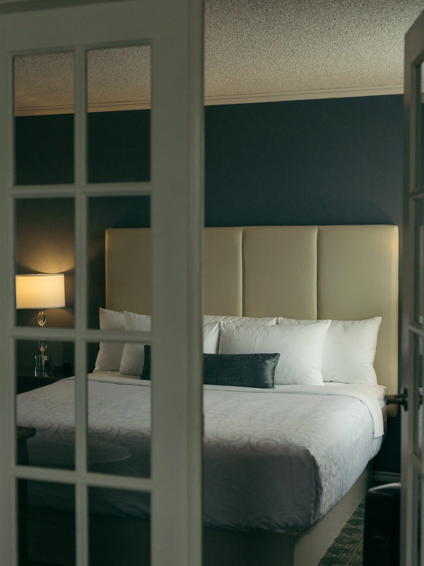 A view of a bedroom through a glass-paneled door. The bed has a white headboard and white bedding, with several pillows including a dark-colored pillow. There are lamps with beige shades on nightstands on either side of the bed, and the walls are pai