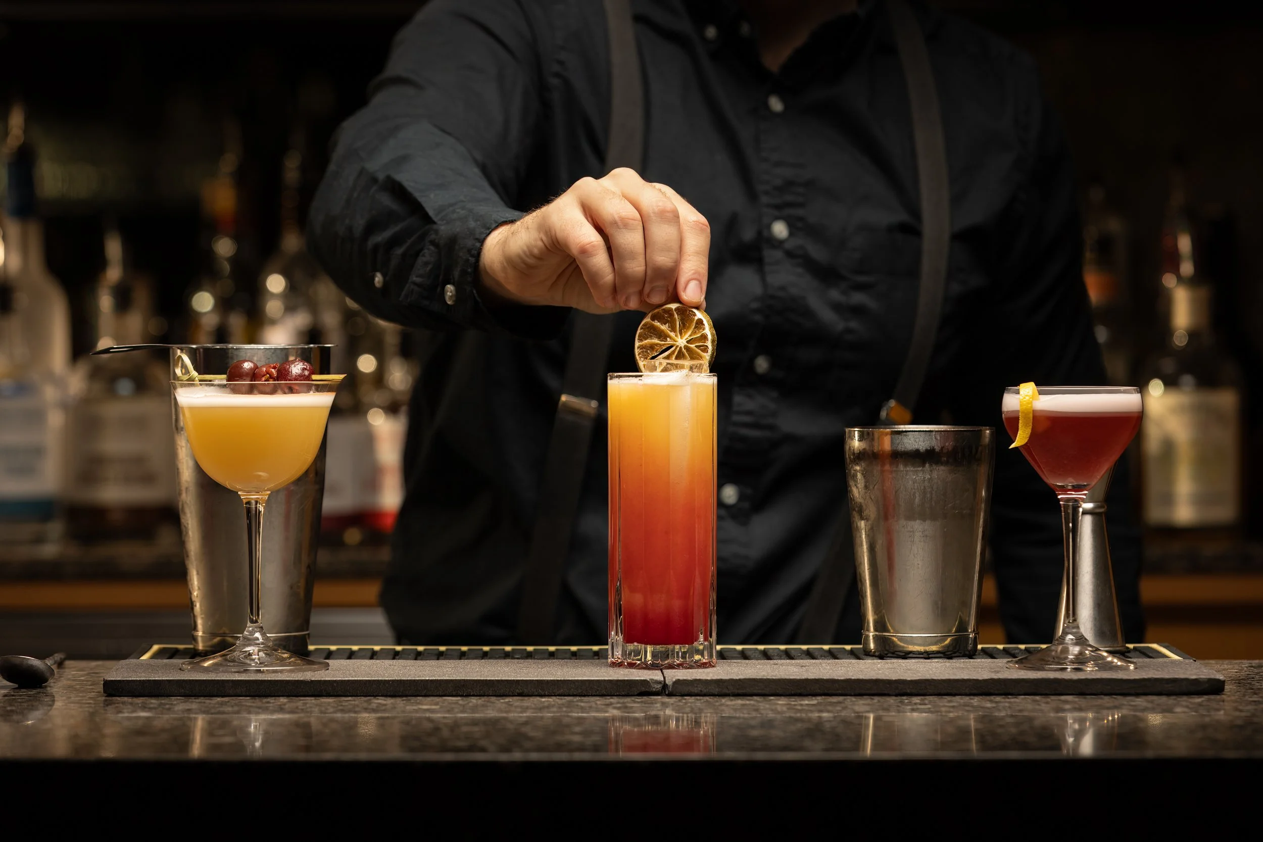 A bartender garnishes a layered cocktail with a dried citrus slice, with two other colorful cocktails and bar tools visible in the background.