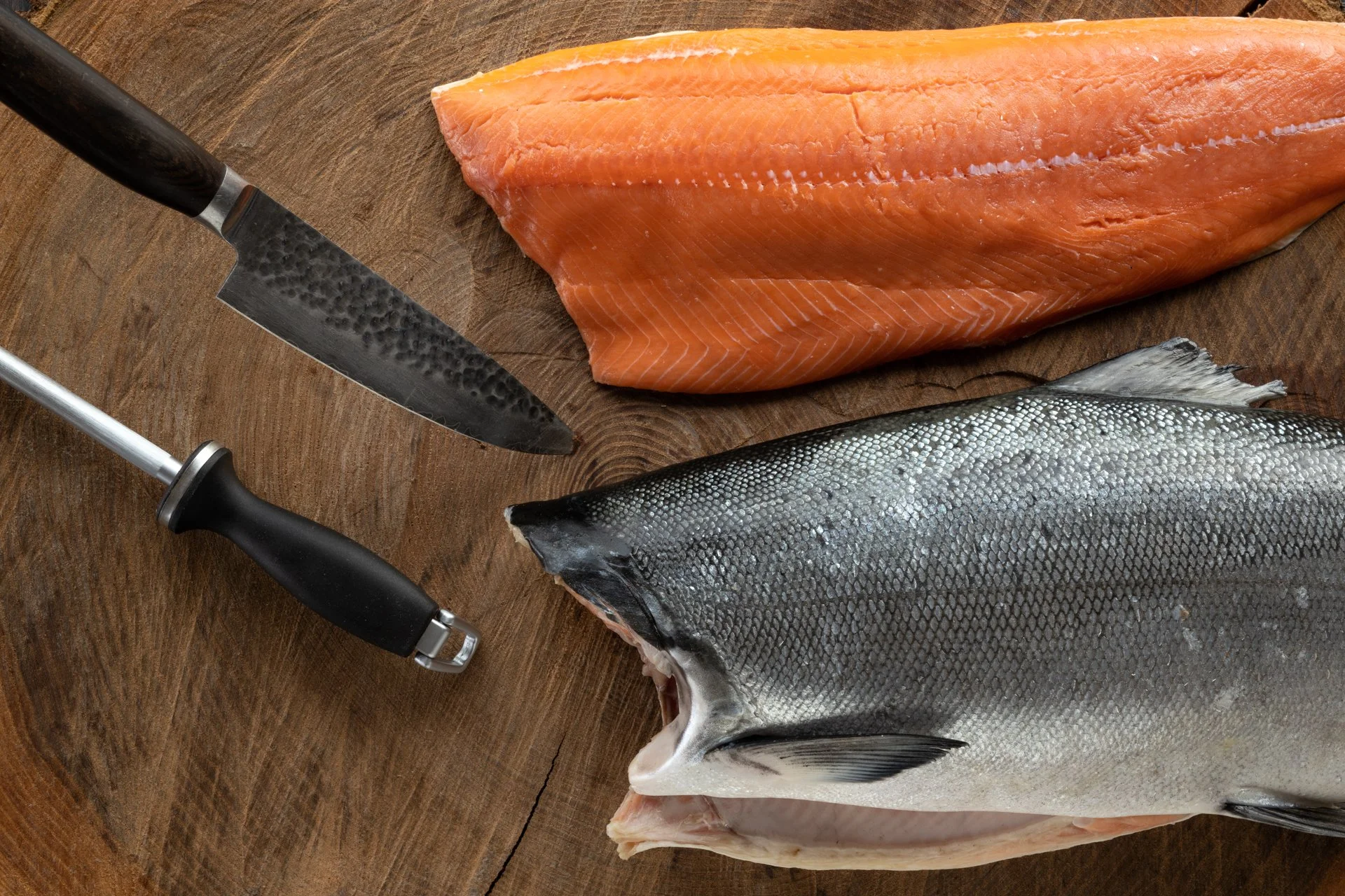 Fresh salmon fillet and whole fish on a wooden cutting board with a filleting knife and a peeler.