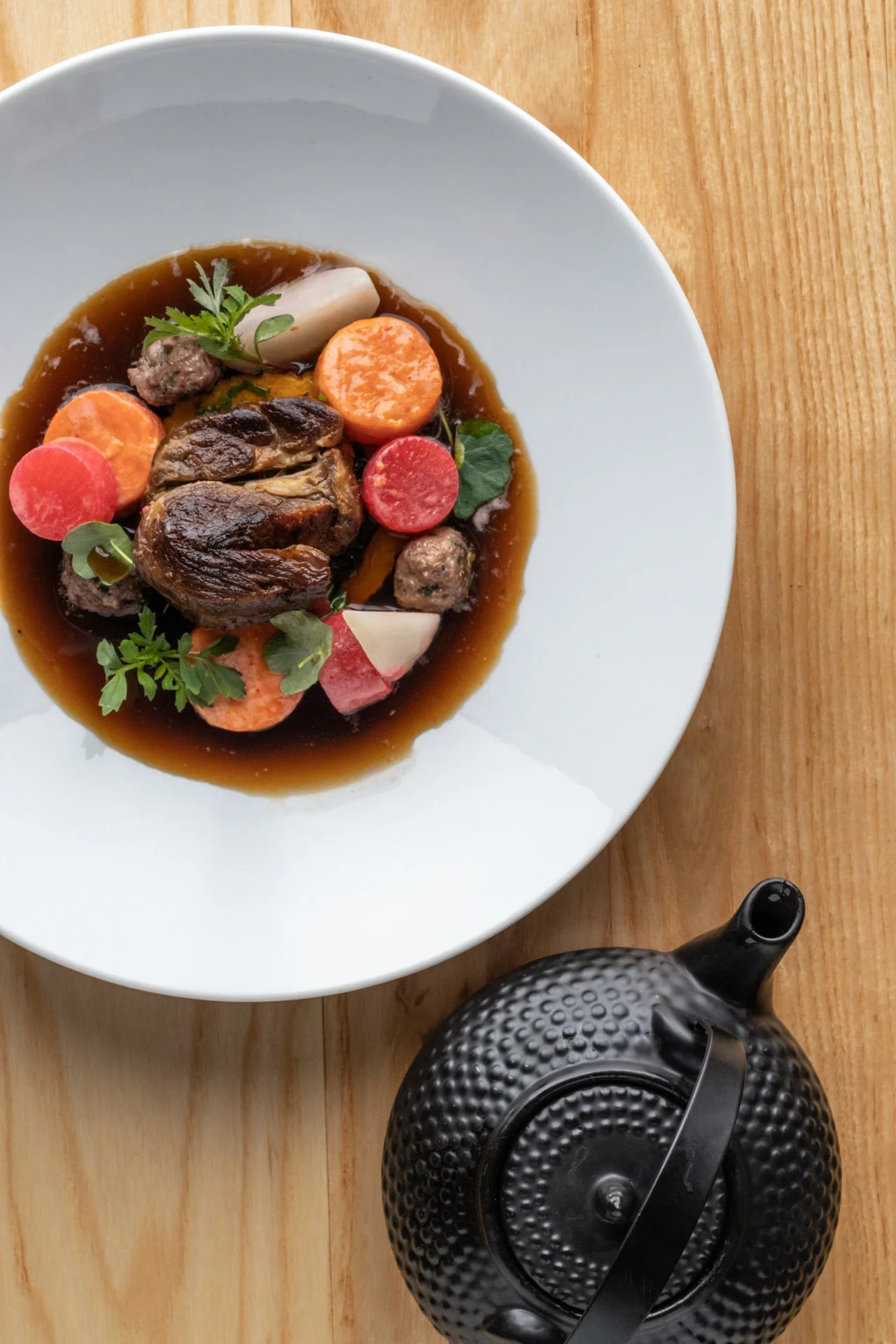 A white bowl of beef stew with carrots, radishes, meatballs, herbs, and a rich brown sauce on a wooden table, with a black teapot nearby.