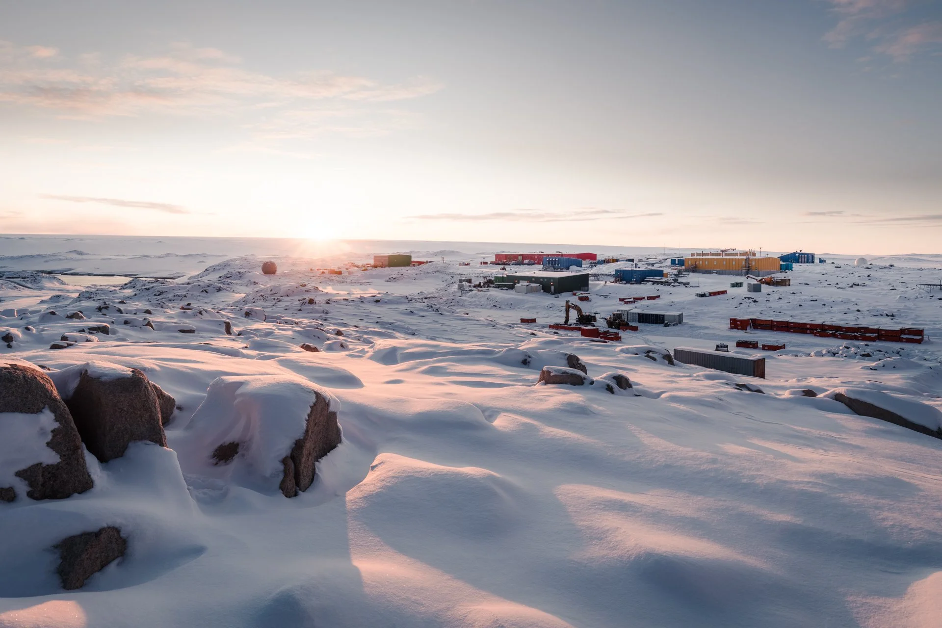 Snow-covered landscape with scattered rocks and a distant chain of colorful buildings under a setting or rising sun, with a clear sky and some clouds.