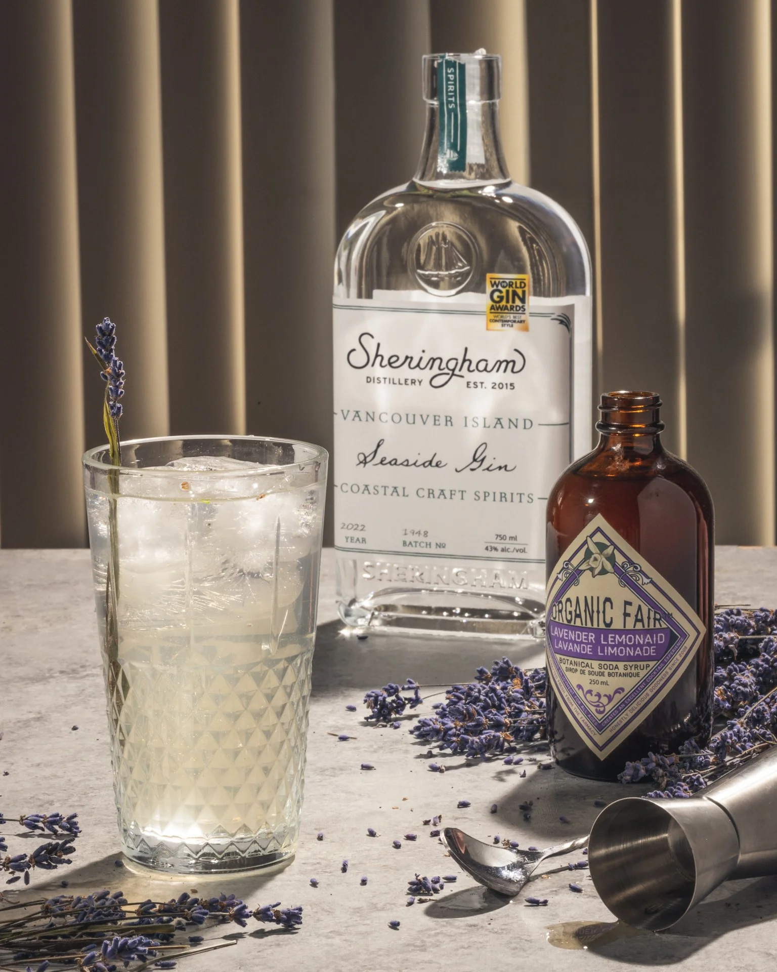 A bottle of Sheringham seaside gin, a glass with a lavender sprig, a bottle of organic lavender lemonade syrup, and a metal cocktail shaker on a light-colored surface with scattered dried lavender flowers and a spoon.