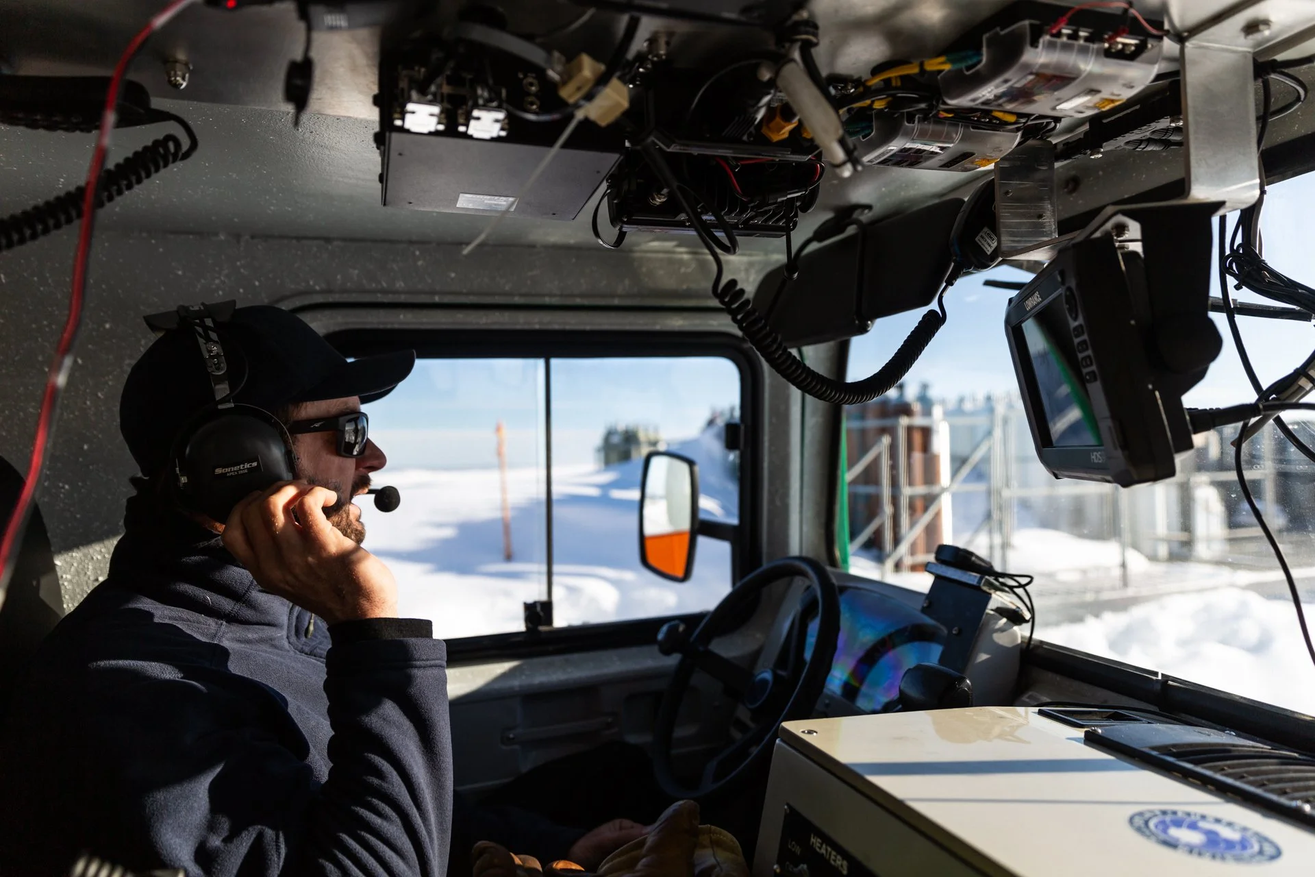 A man wearing sunglasses and a headset talking on a radio inside a vehicle, with various electronic equipment mounted above him and a snowy outdoor scene visible through the windows.