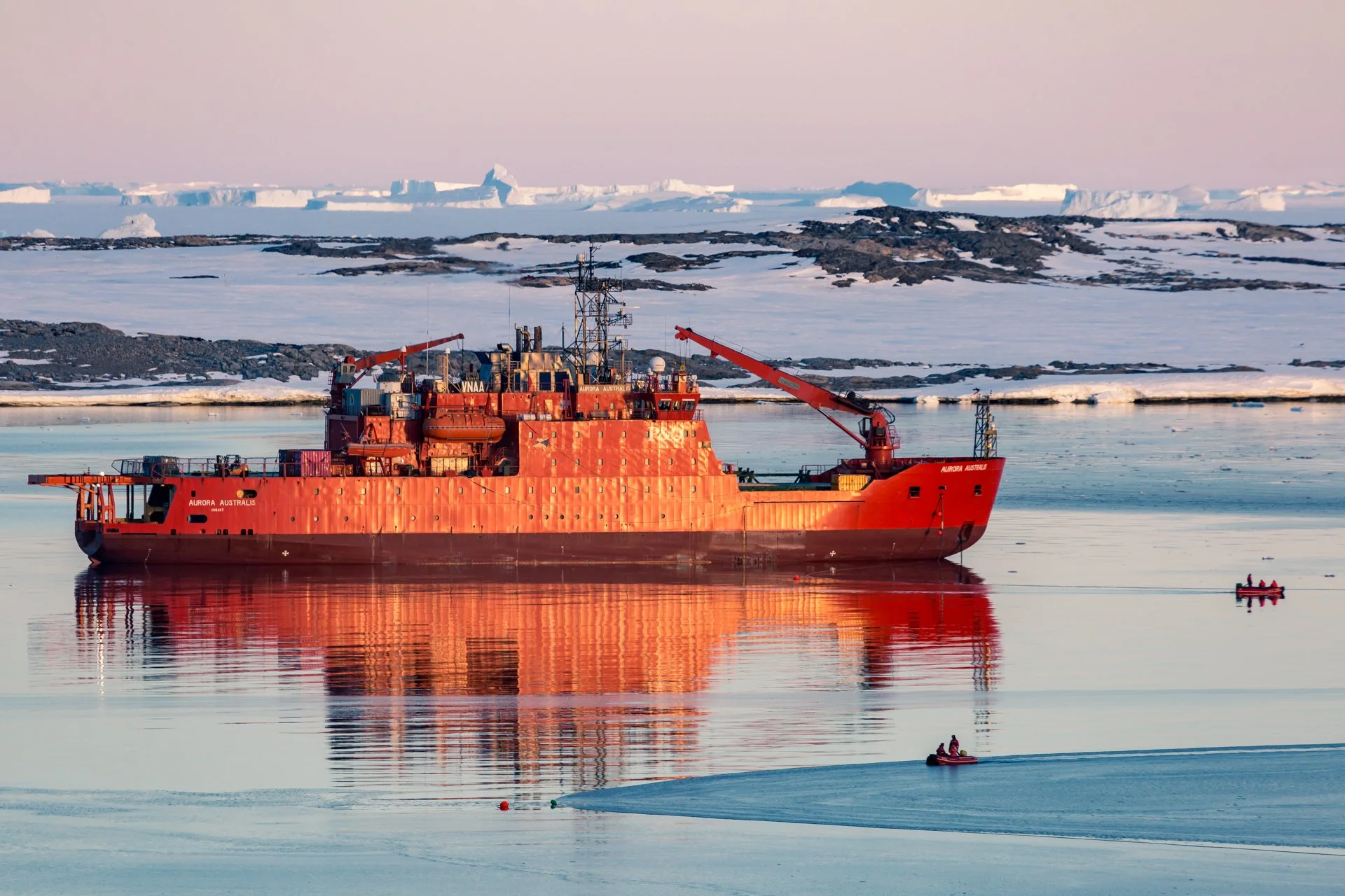A red icebreaker ship named Aurora Australis sailing in icy waters with snow-covered land and glaciers in the background.