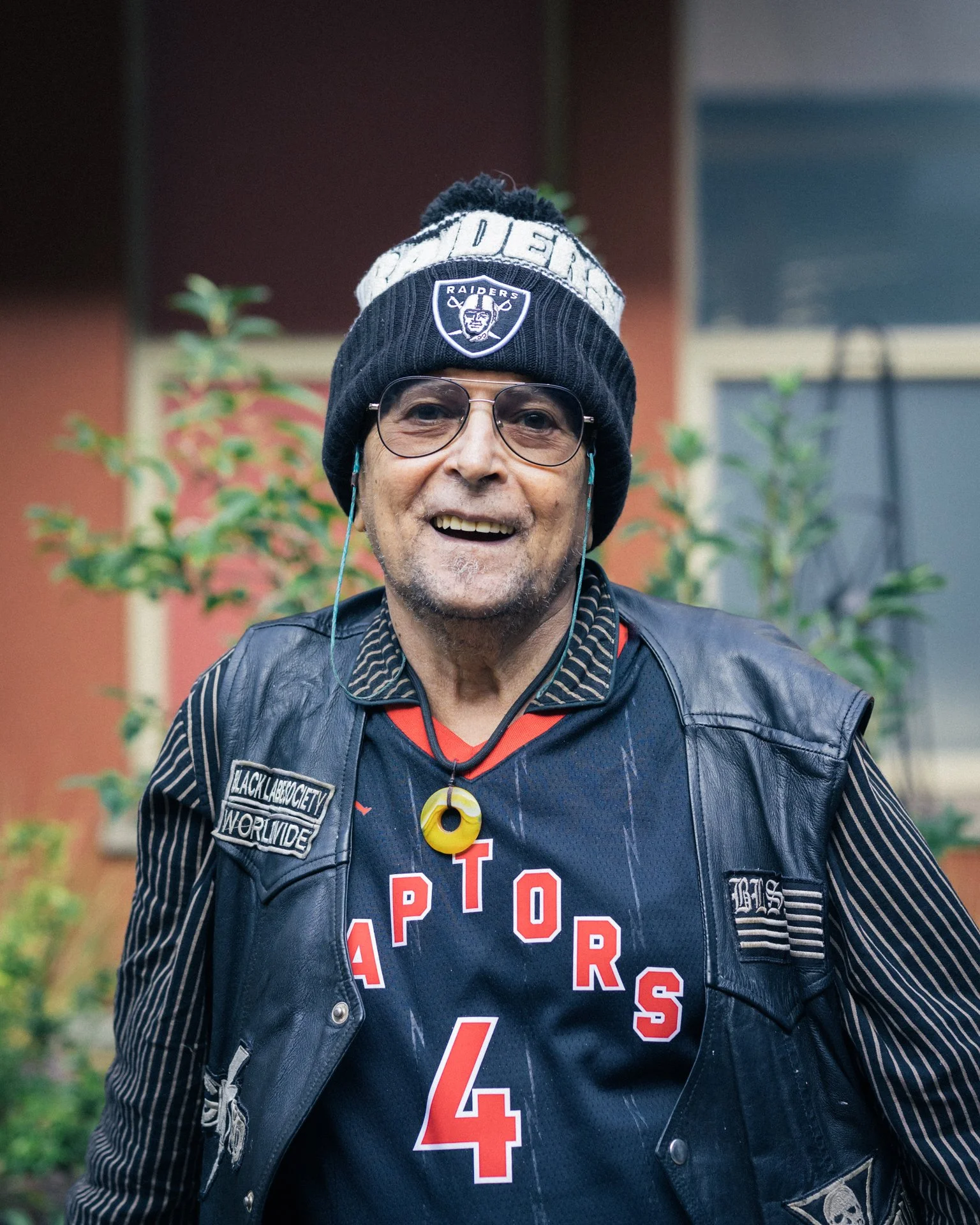 A man wearing glasses and a black beanie with the Raiders logo, smiling, dressed in a black leather vest over a sports jersey, outdoors with greenery in the background.
