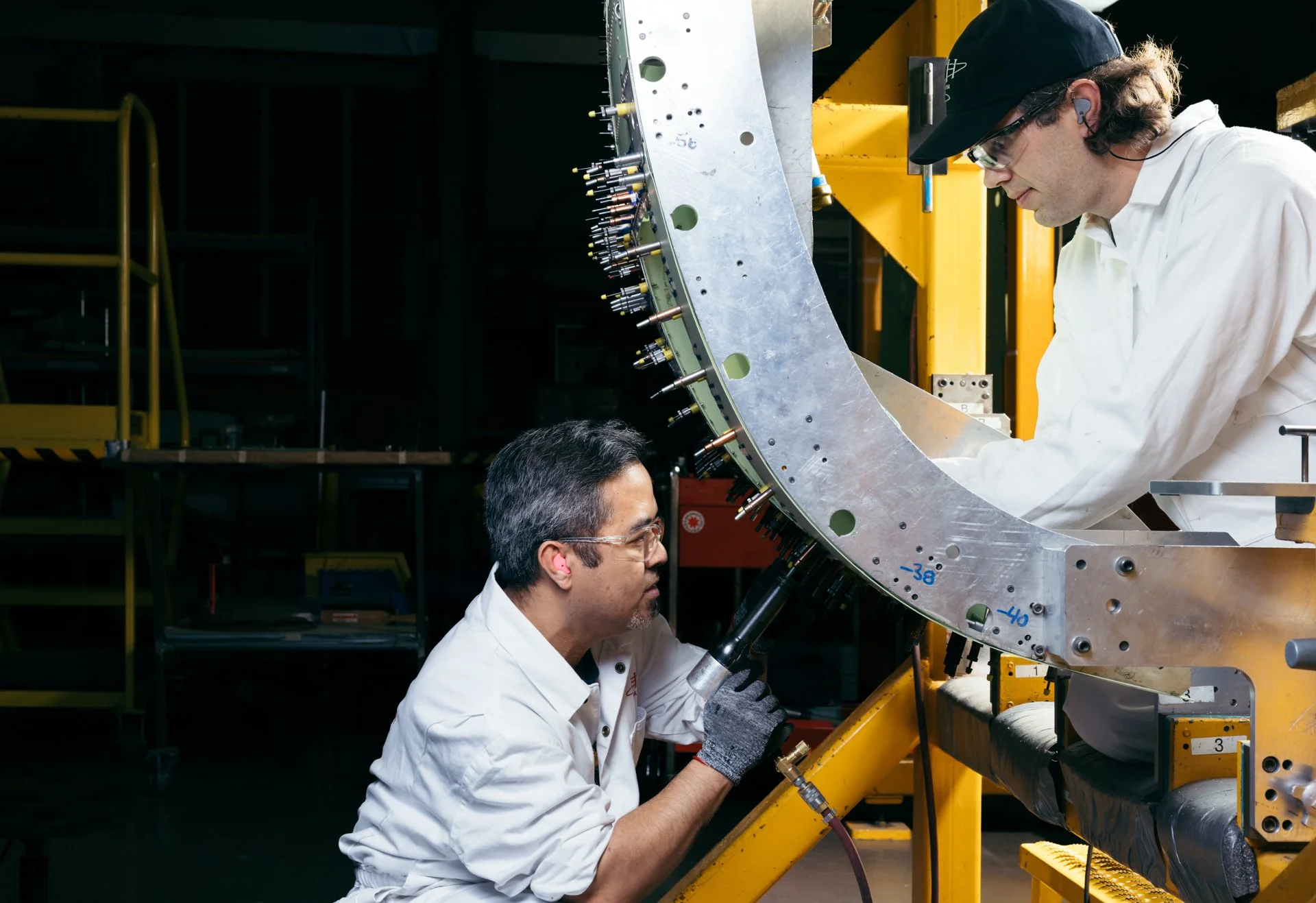 Two engineers working on a large metallic spacecraft component in an industrial setting. One is kneeling and the other is standing, both wearing white lab coats. The component is mounted on a yellow frame, with tools and pipes visible around them.