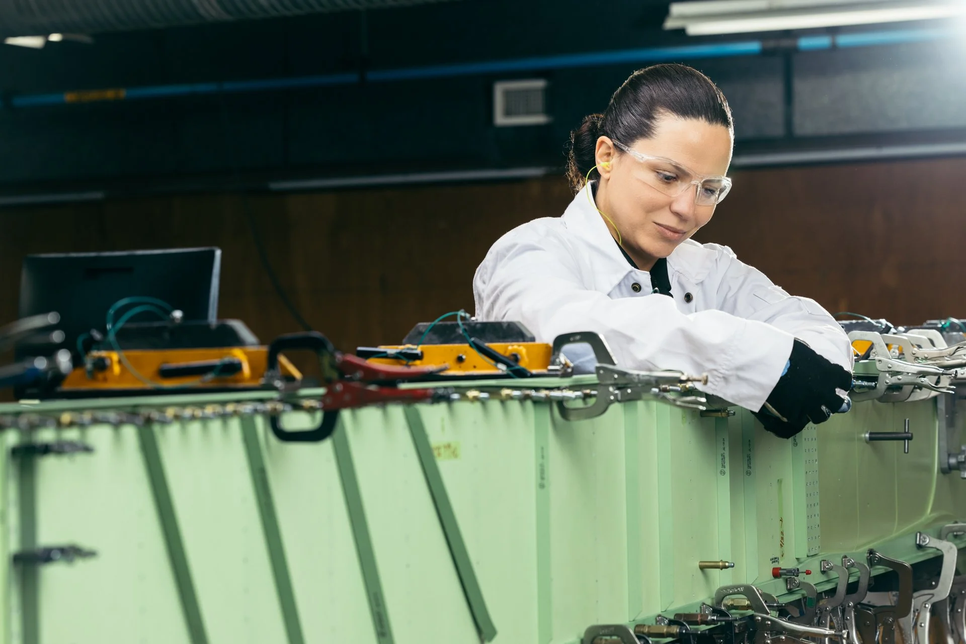 A woman in safety glasses and a white lab coat working with machinery at an industrial or research facility.