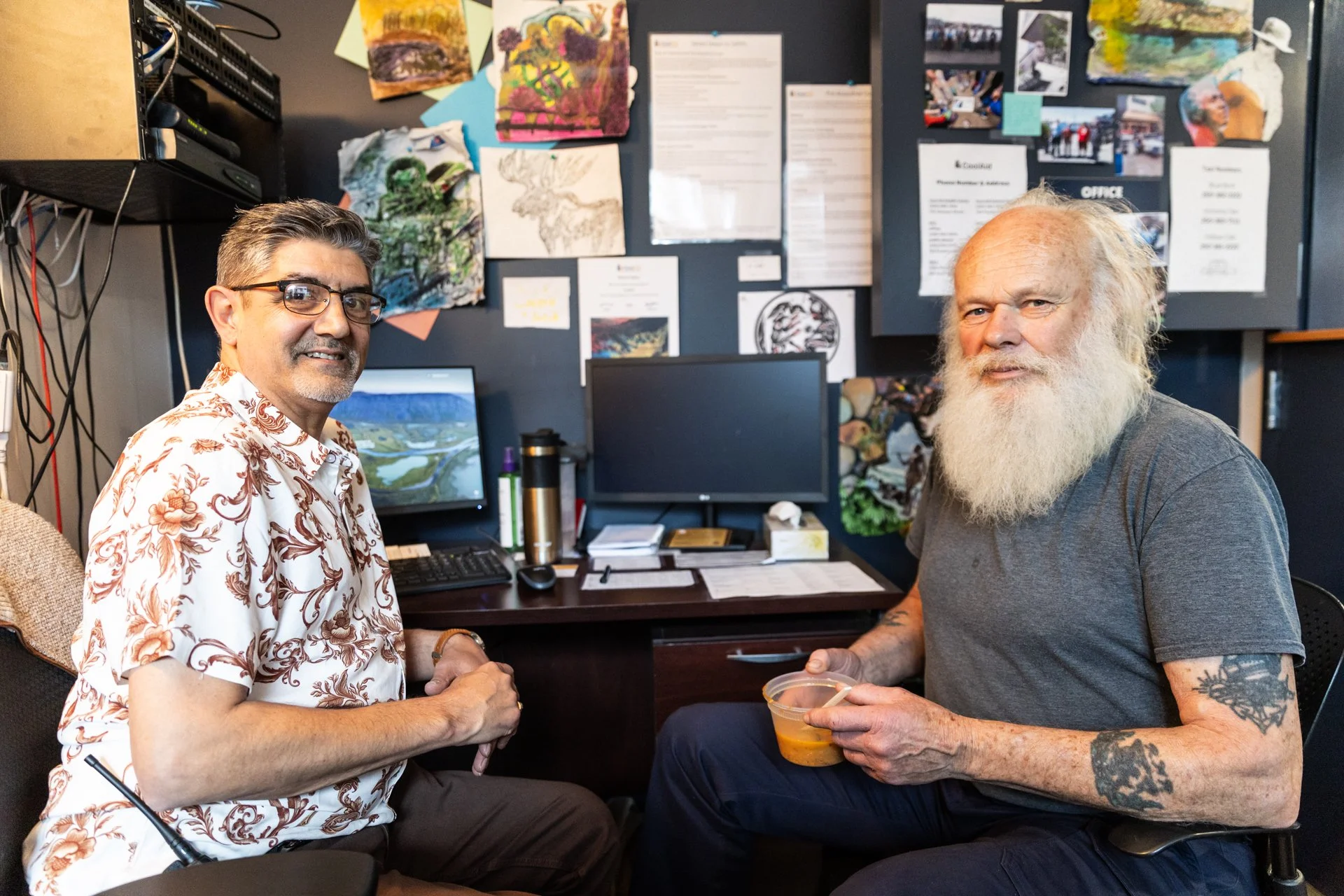 Two men shaking hands in an office, with art and photos on the wall behind them and computer monitors on the desk.