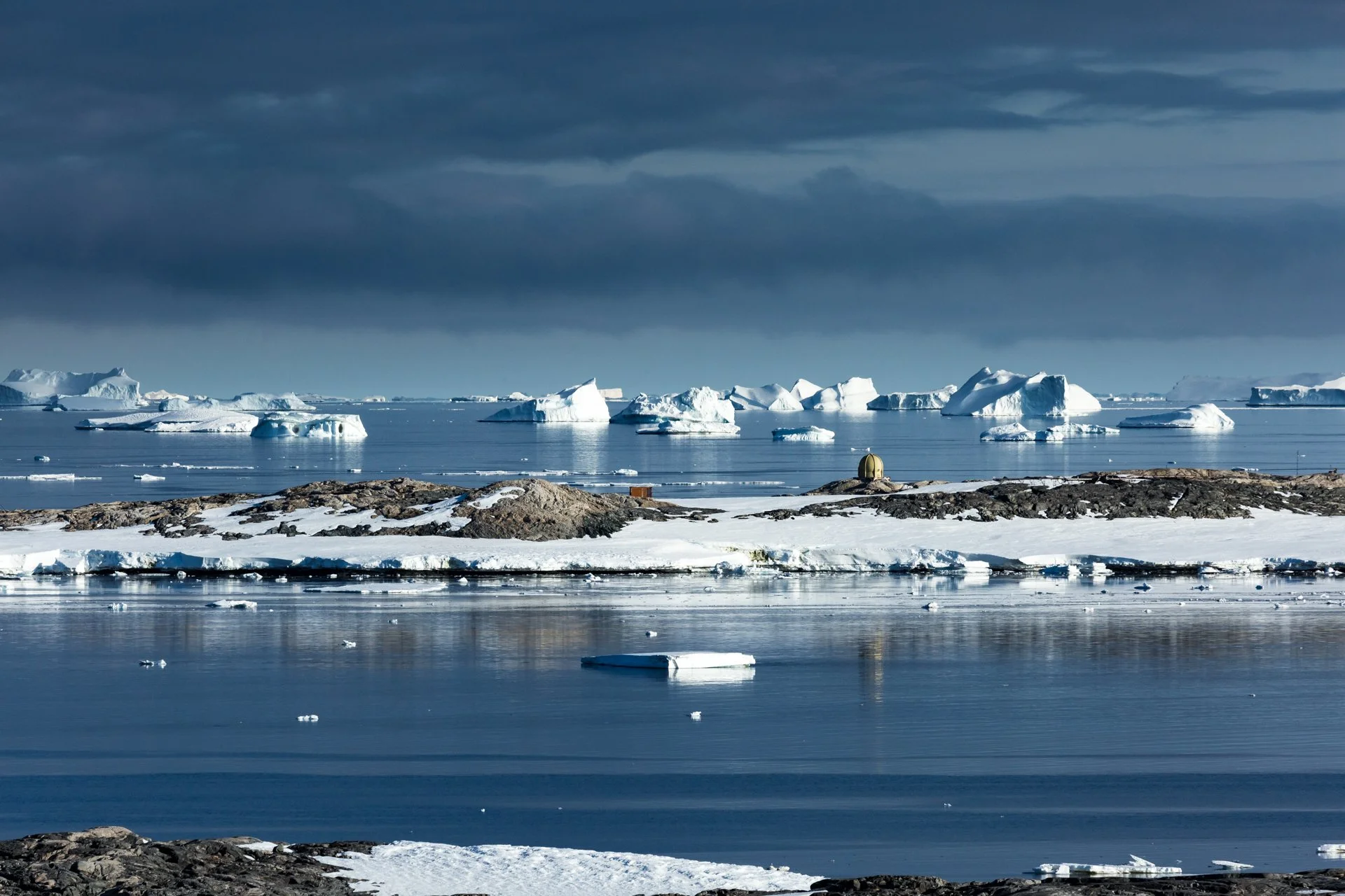 Icebergs floating in a calm Arctic or Antarctic ocean with snow-covered rocks in the foreground and dark clouds overhead.