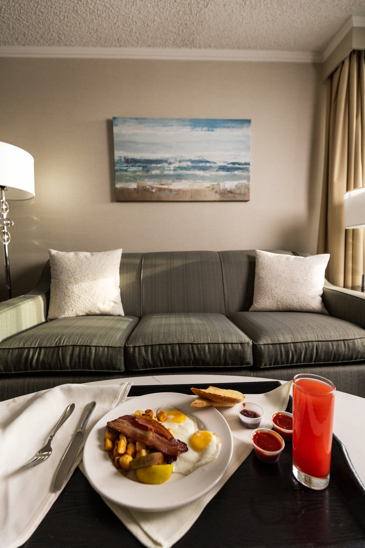 Breakfast with eggs, bacon, toast, ketchup, and juice on a tray, in front of a living room with a gray sofa, white pillows, a painting, and beige curtains.