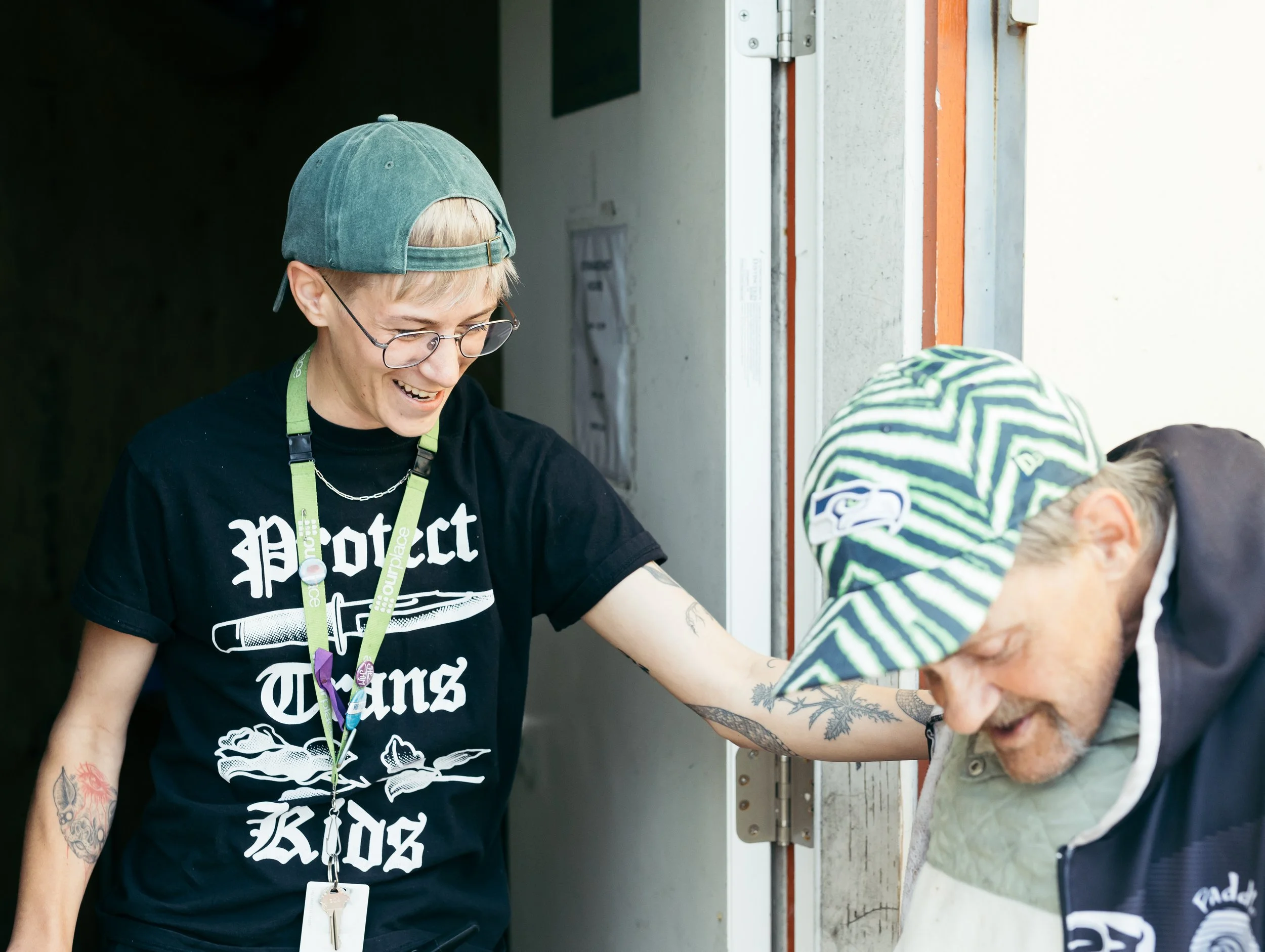 A person wearing glasses and a green cap, smiling and helping an elderly man with tattoos on his arms, who is bending down, in front of a door frame.