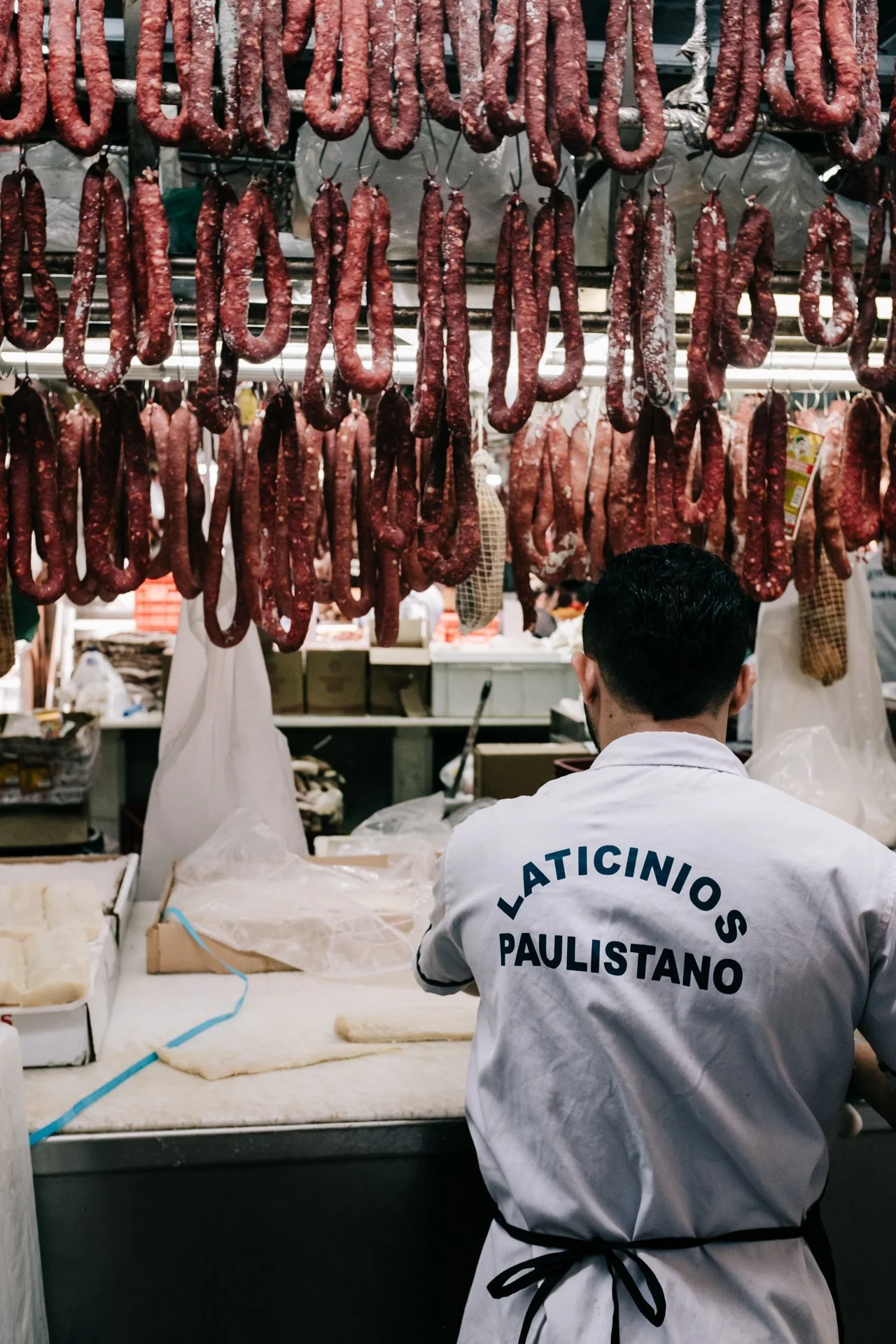 A butcher shop with sausages hanging from hooks, and a worker wearing a white apron with 'LATINO PAULISTANO' on the back, seen from behind.