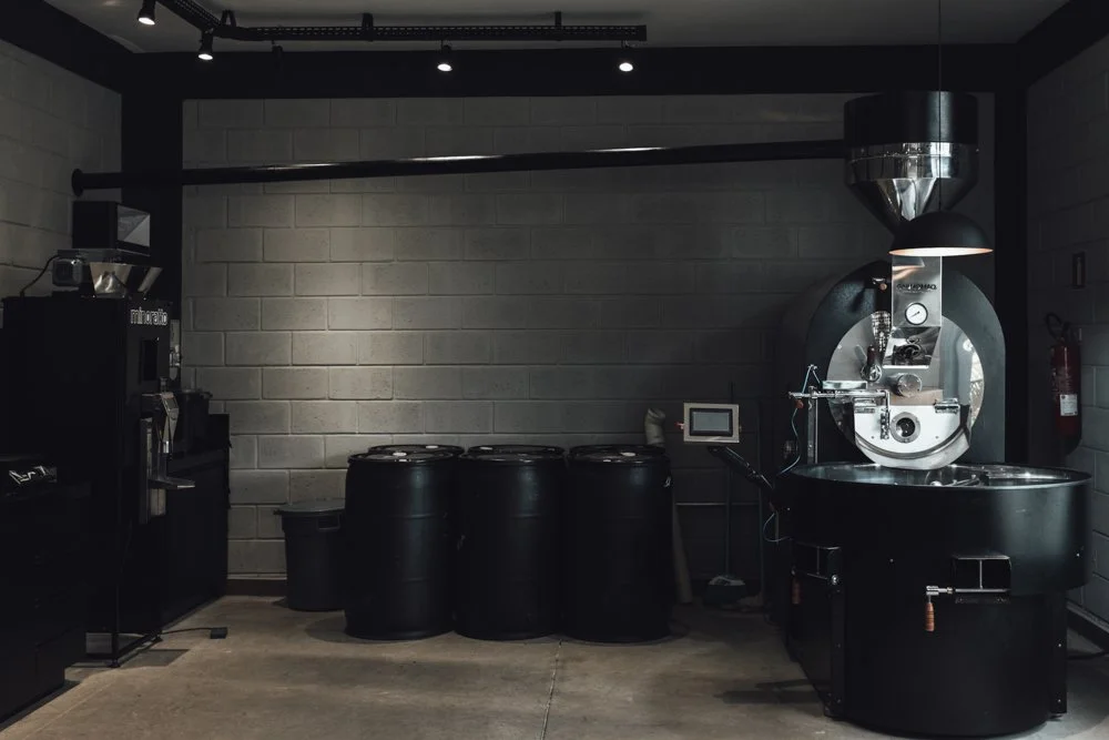 Coffee roasting equipment, including a roaster machine, several barrels, and a coffee grinder set up in a modern industrial style space with concrete floors and brick walls.