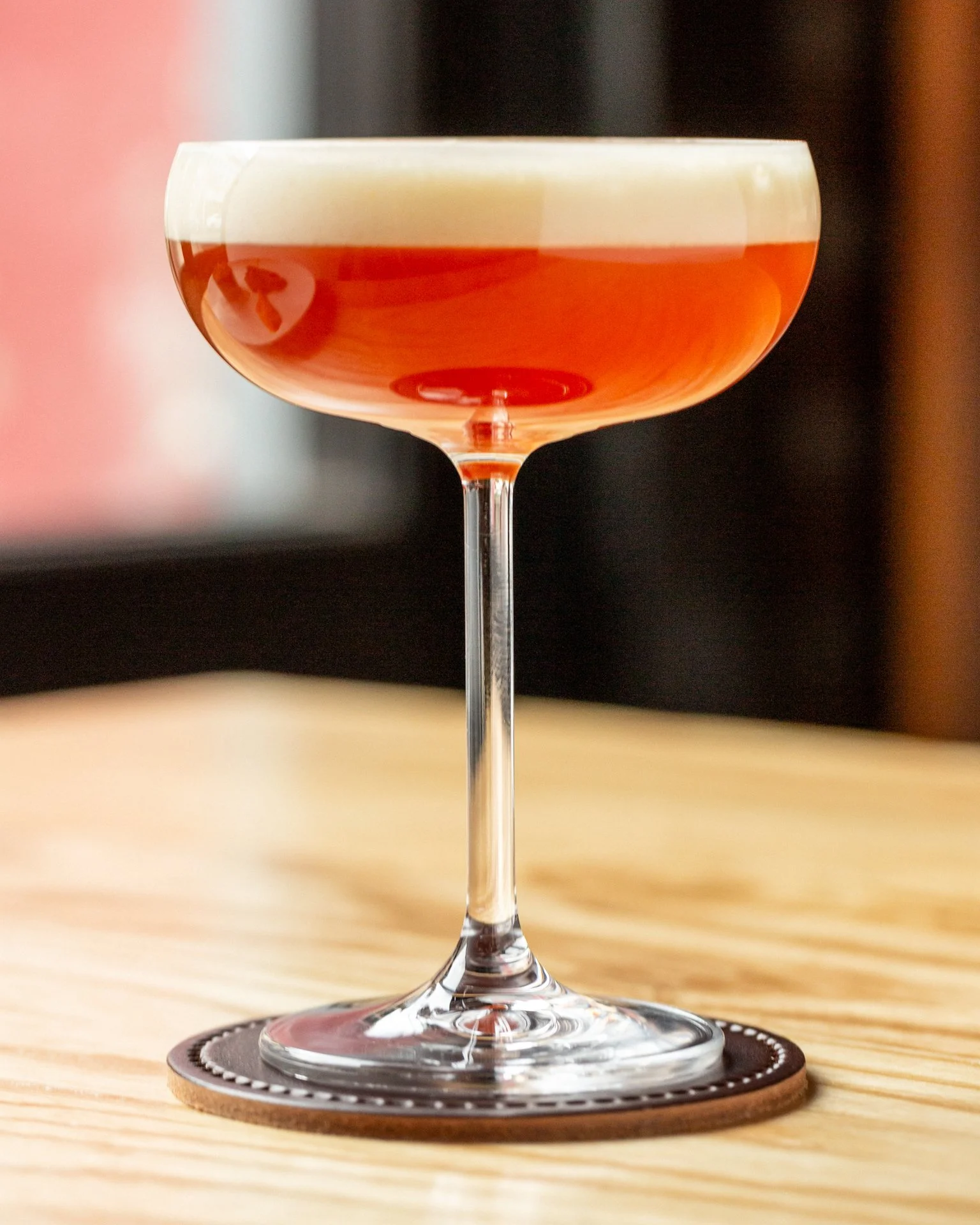 Close-up of a cocktail glass filled with an orange-colored beverage, topped with a foamy white layer, placed on a wooden table with a leather coaster underneath.