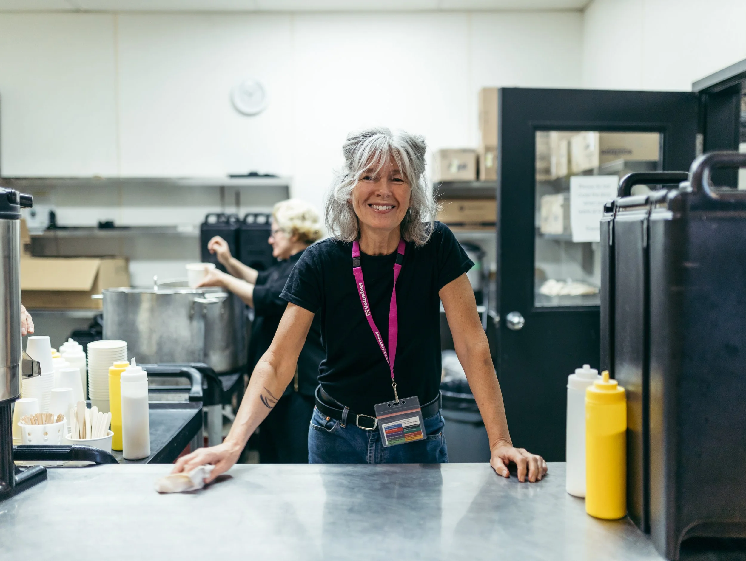 A smiling woman in a black shirt and jeans cleaning a countertop in a kitchen or cafeteria setting, with condiment bottles and cups in the foreground and another woman in the background.