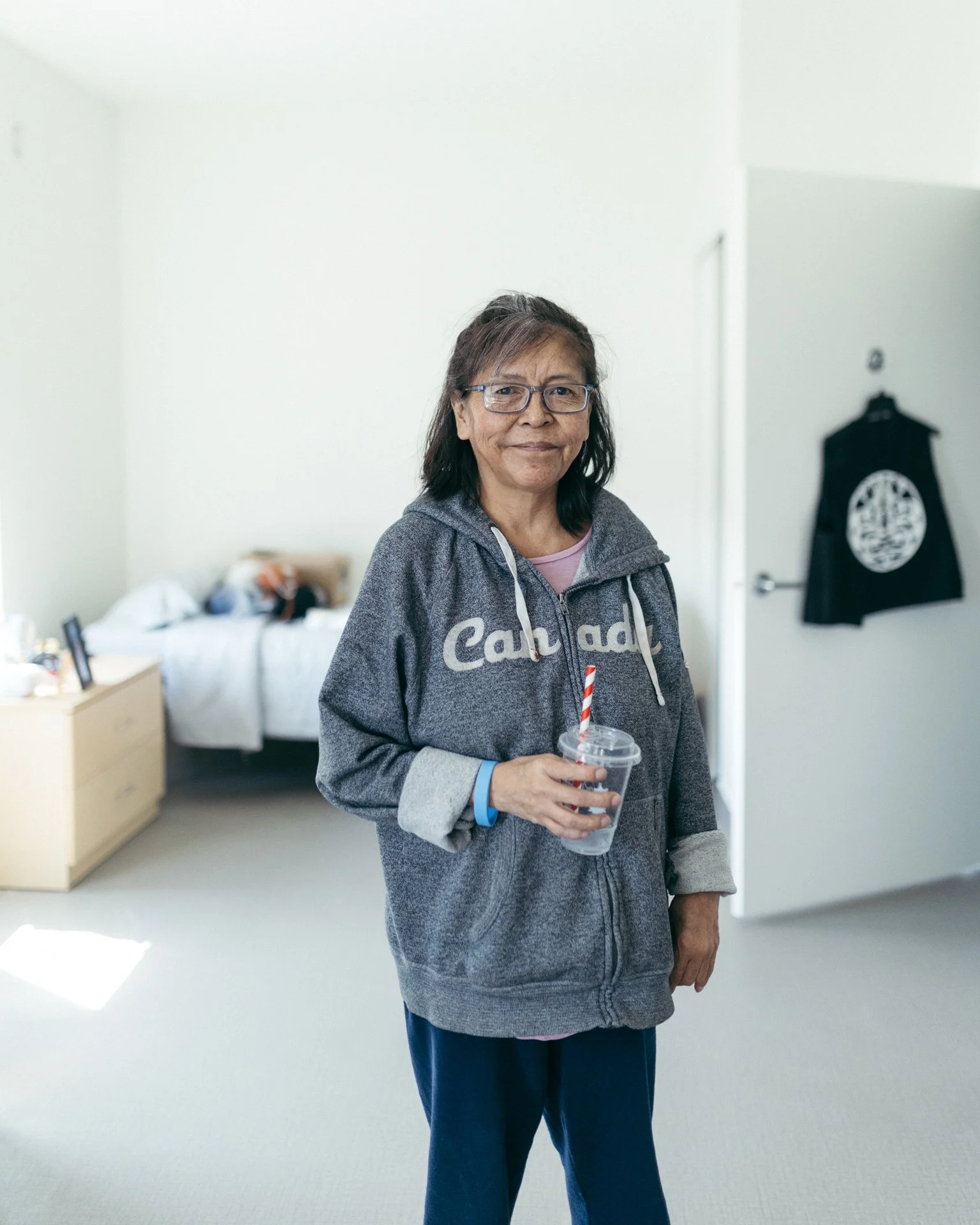 A woman with glasses smiling and holding a clear cup with a striped straw, standing in a room with a bed and a black vest with a white logo hanging on the wall in the background.
