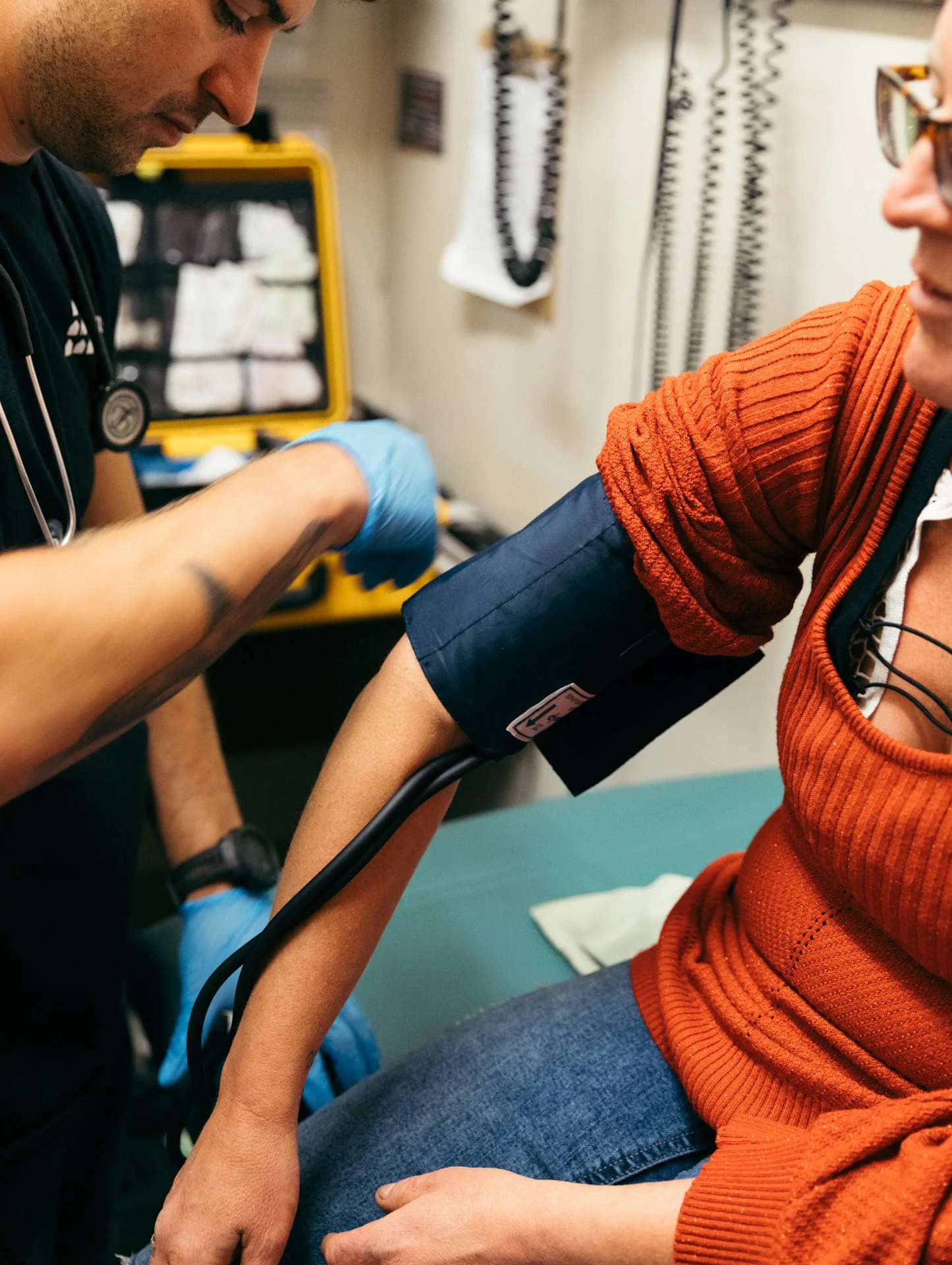 A person wearing an orange sweater is getting a blood pressure check from a healthcare professional. The professional is wearing gloves and using a blood pressure cuff on the person's arm. The background includes medical equipment and supplies.
