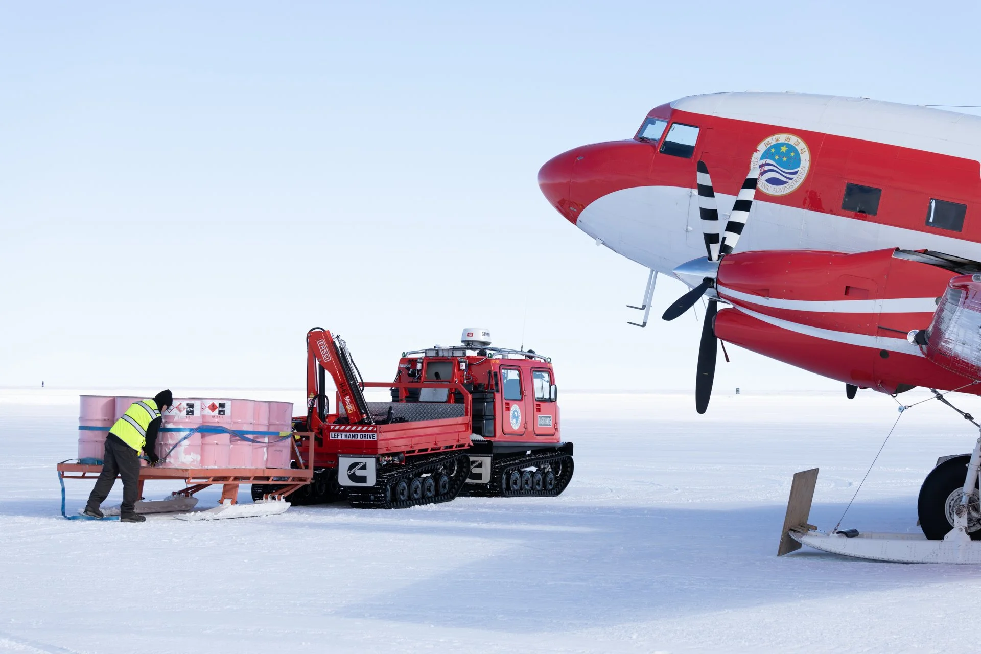 A person in a yellow safety vest working near a large pink container in a snowy landscape, with an airplane and ground support equipment in the background.