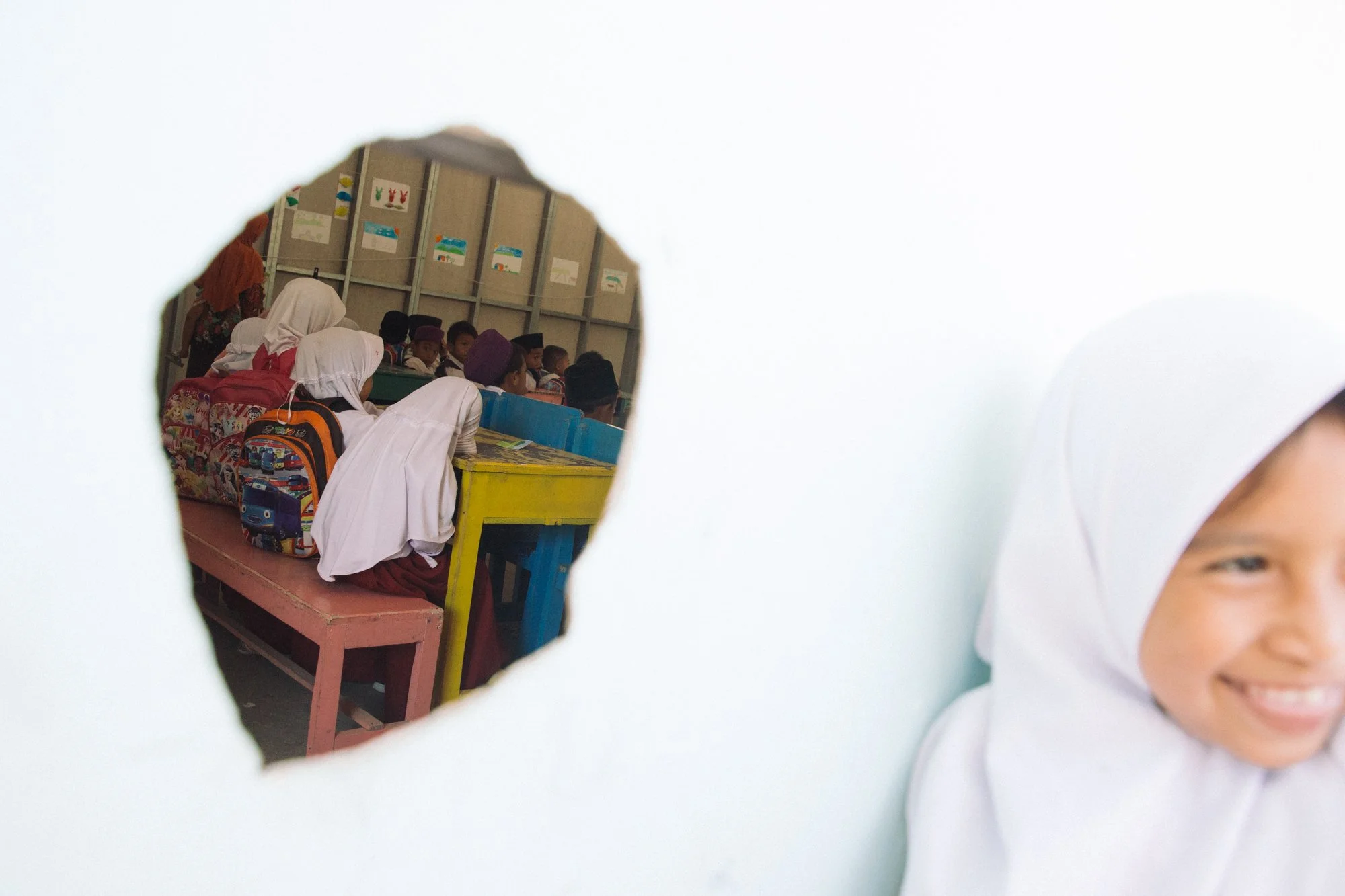 Children wearing white and colored clothes and hats sitting at desks inside a classroom, view seen through a hole in a white wall.