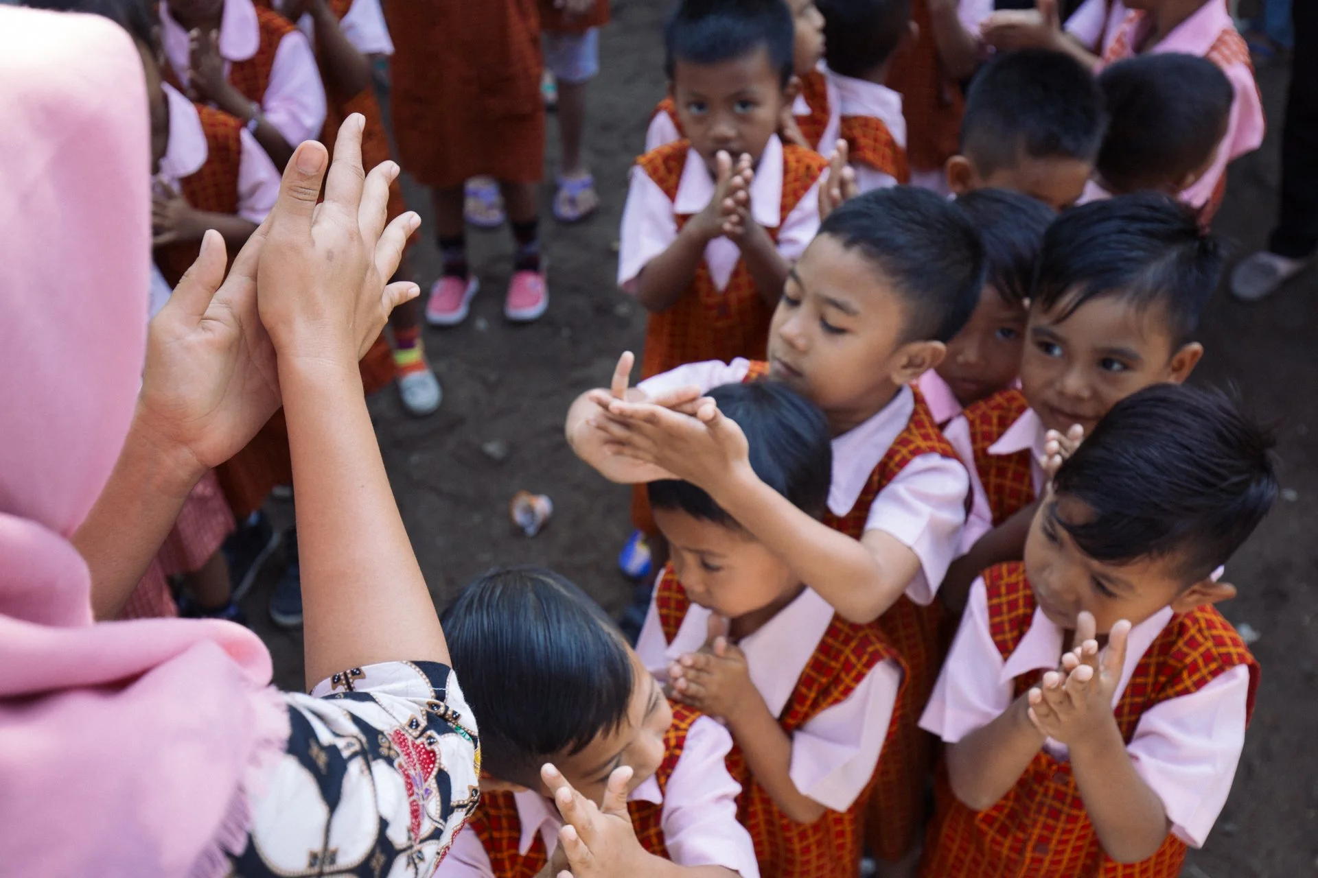 A group of school children in uniforms, some clapping and others with their hands folded, gathered around a person wearing a pink headscarf who is holding their hands up for a hands-on activity or demonstration.