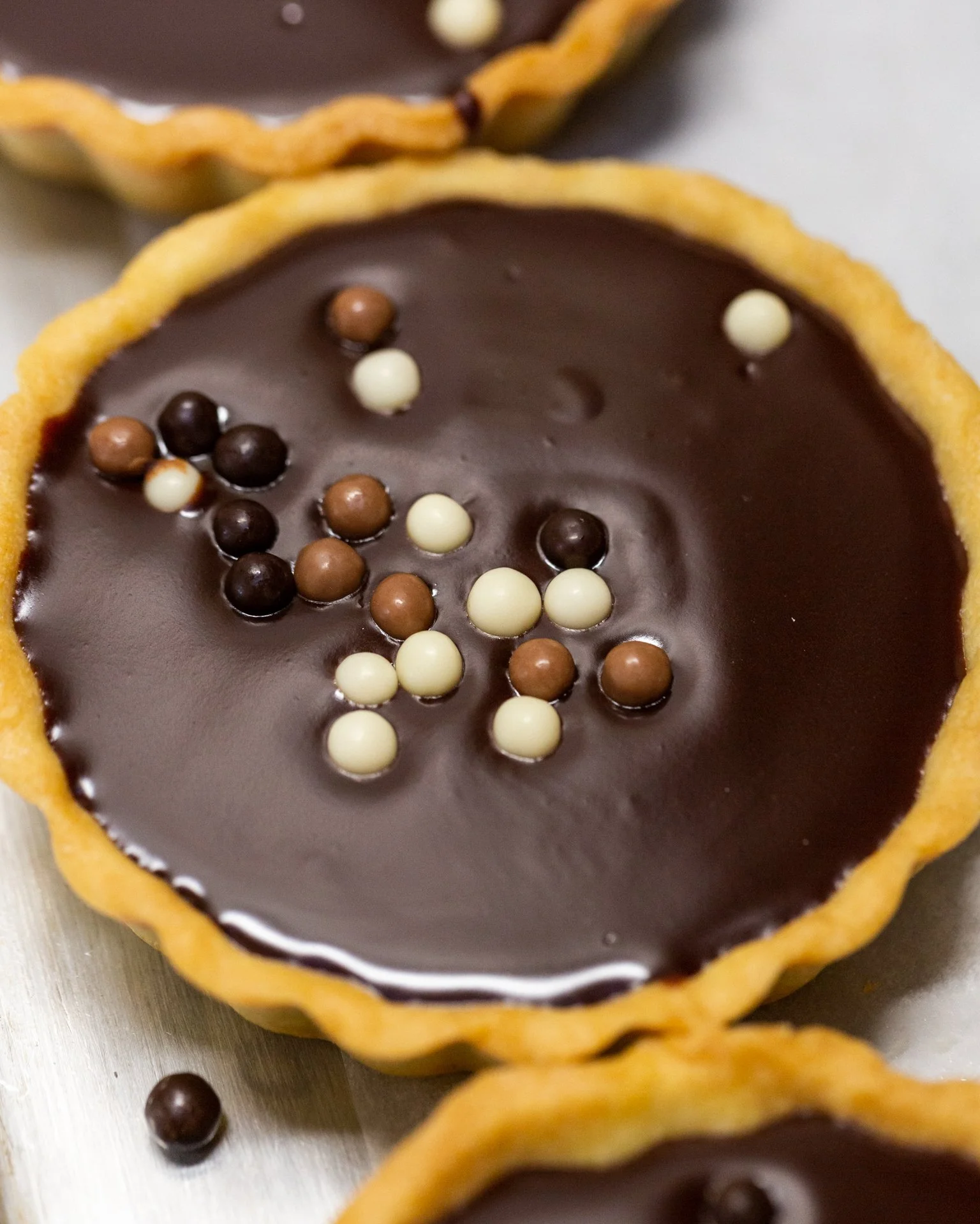 Close-up of chocolate tartlets with a golden crust, filled with smooth chocolate ganache, topped with small white, light brown, and dark brown spherical chocolate decorations.