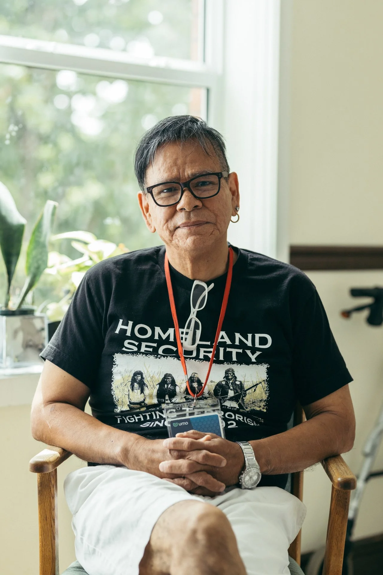 A man with glasses sitting on a wooden chair indoors, wearing a black 'Home and Security' T-shirt, holding a smartphone, with a window and potted plants behind him.