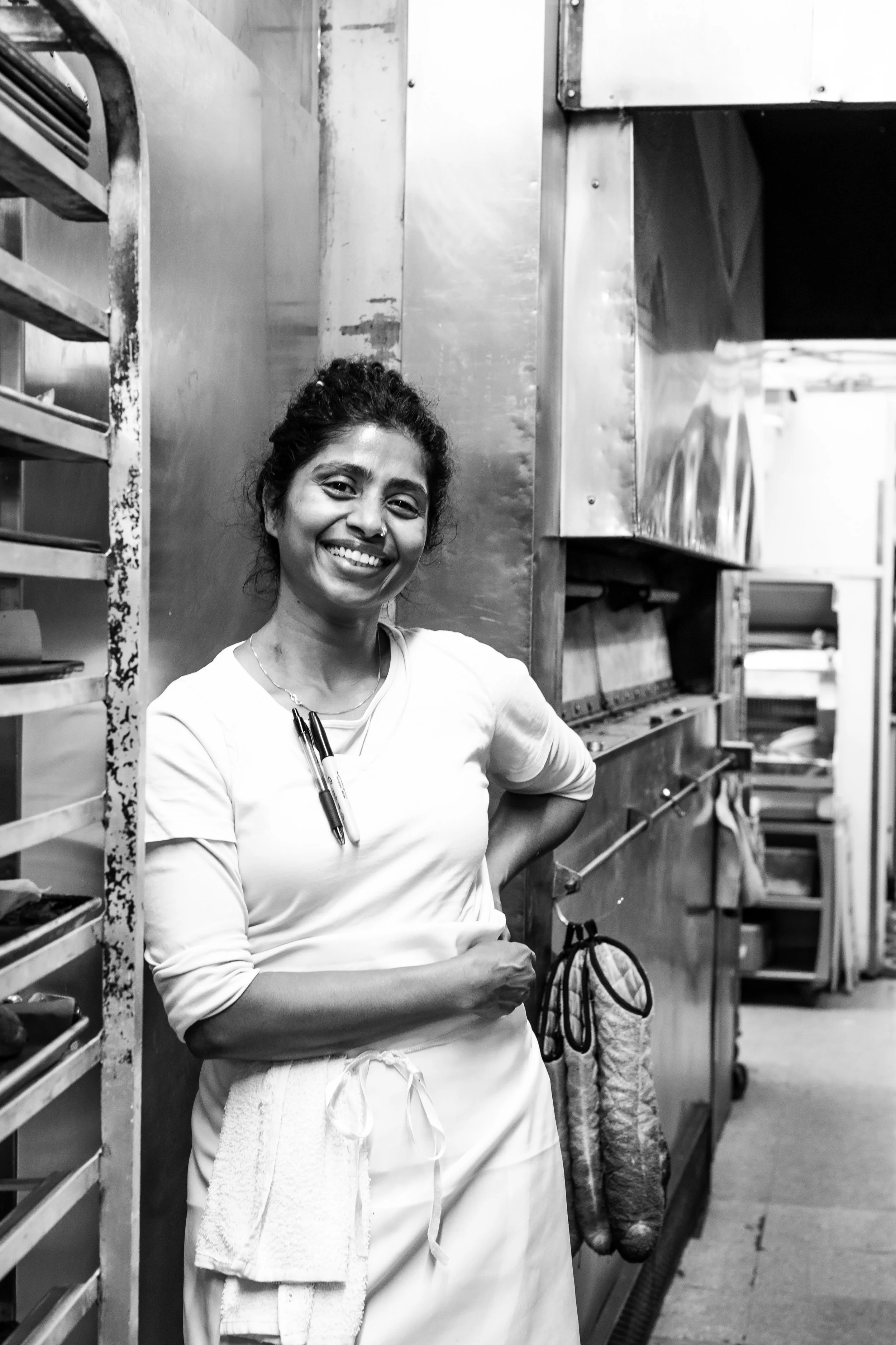 A smiling woman with dark curly hair tied back, wearing a white long-sleeve shirt, apron, and holding a marker, stands in a kitchen next to metal shelves with kitchen equipment and utensils.