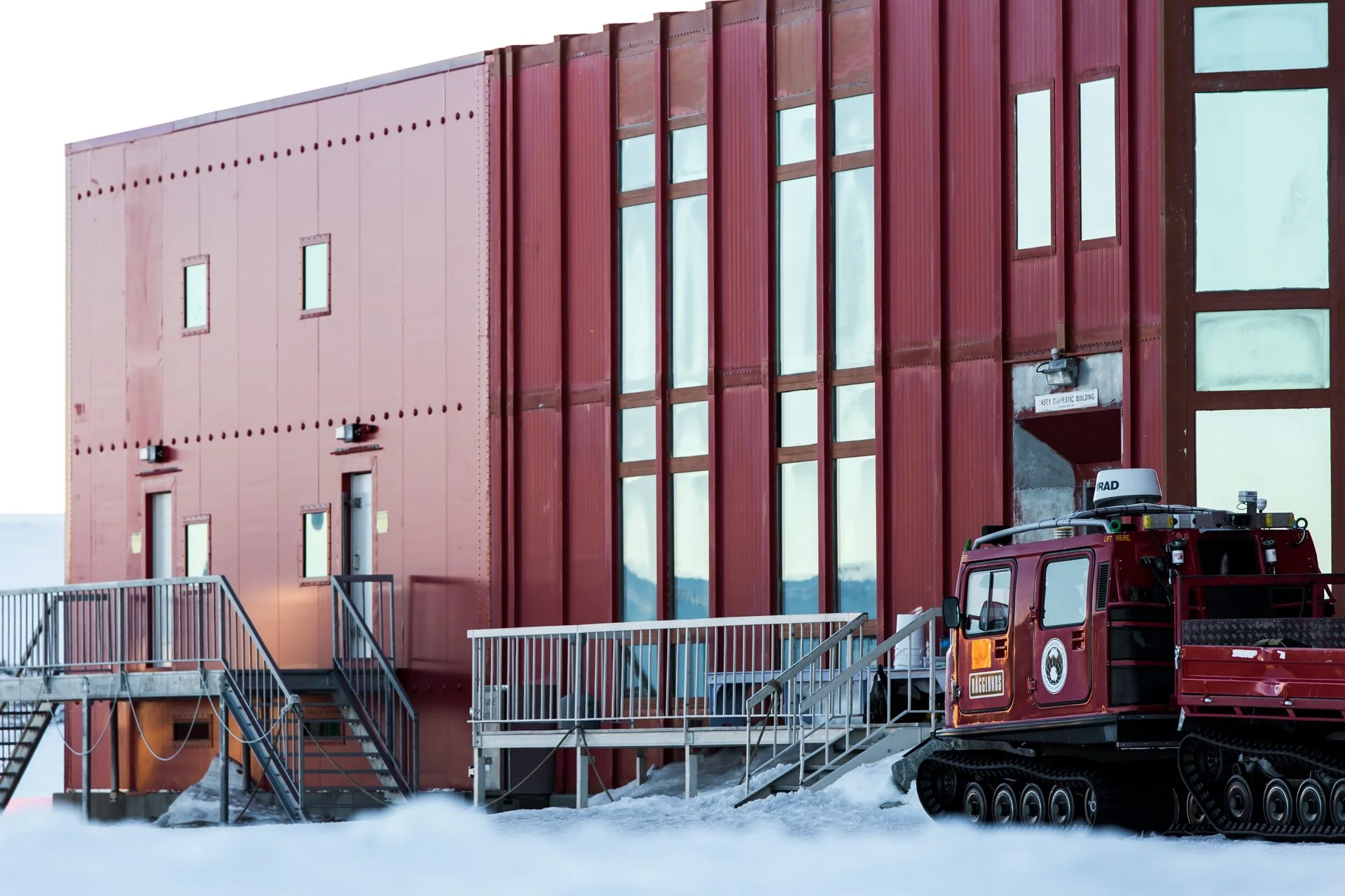 A red building with a modern design and large glass windows, part of the building has a sign that reads 'Casey Dulcic Building'. Snow is on the ground, and a red tracked fire truck is parked nearby, with equipment on the roof.