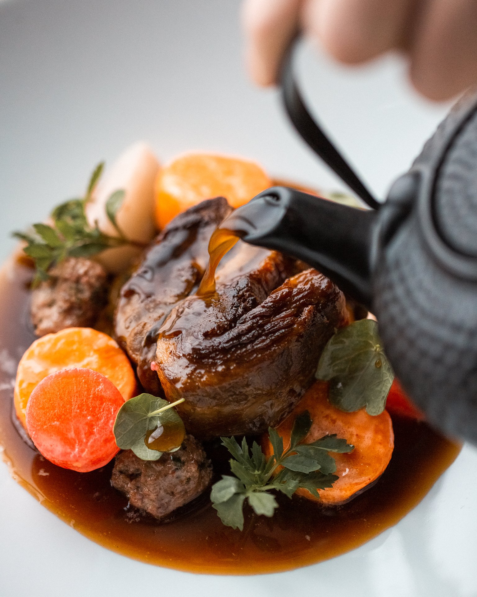 A close-up of a plated meal featuring a large piece of cooked meat, sliced carrots, herbs, and sauce.