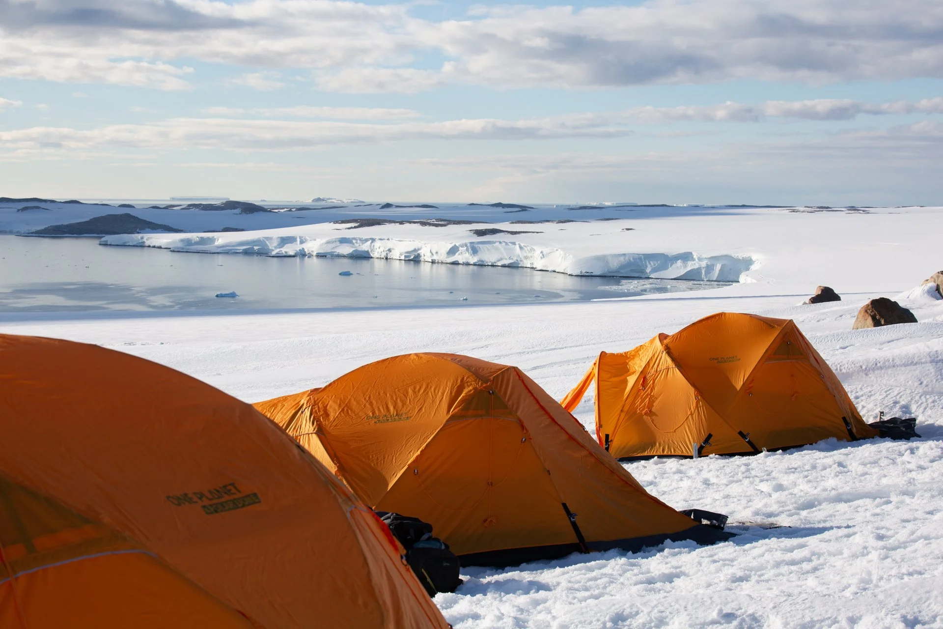 A row of orange tents on snow, with a backdrop of icy water, glaciers, and snowy hills under a cloudy sky.