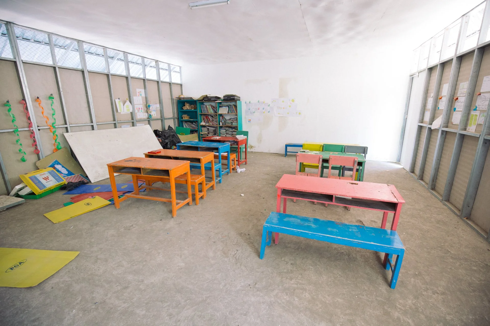 Unfinished classroom with colorful desks and chairs, bookshelves, and children's artwork on the wall.