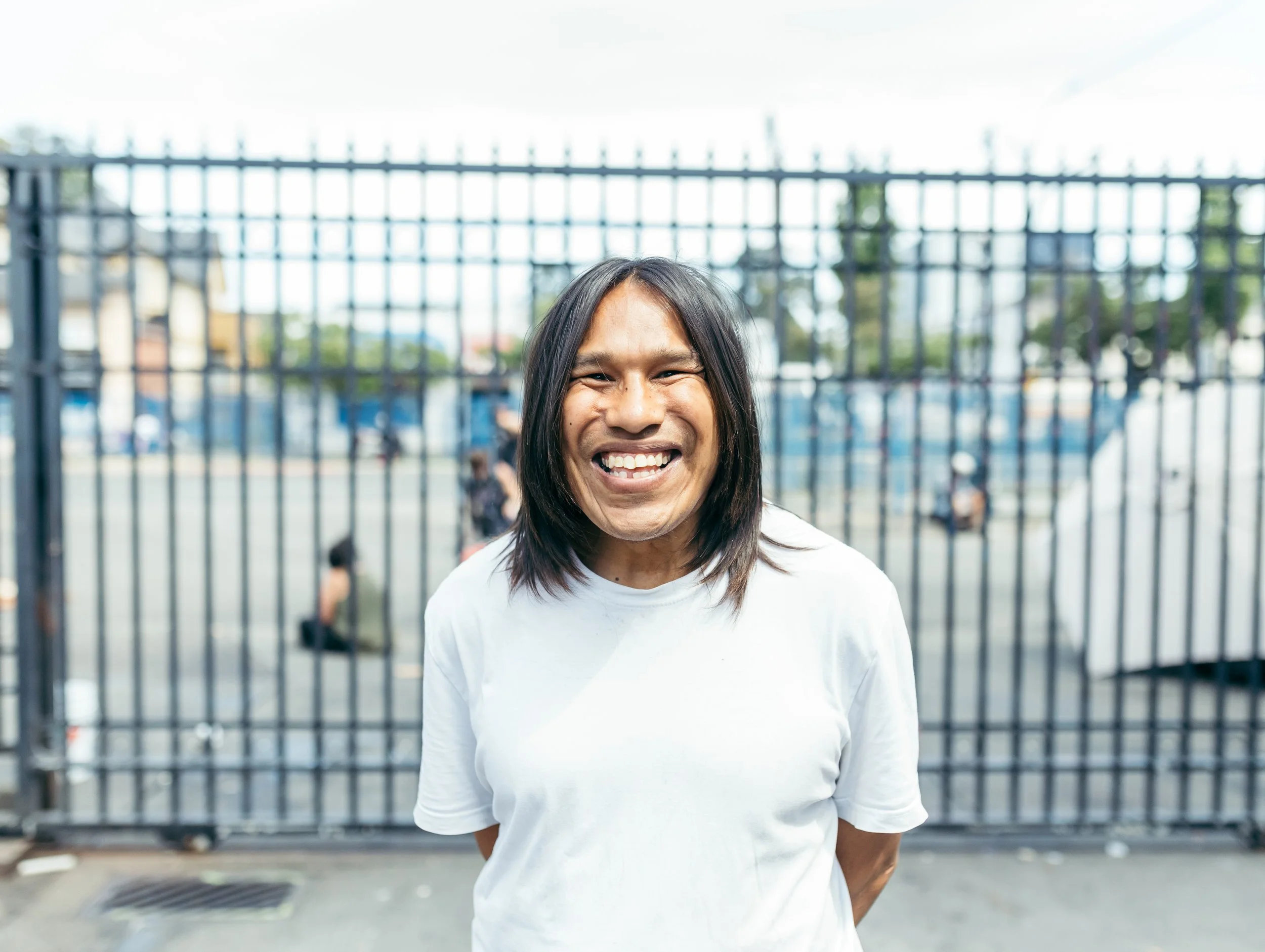 A smiling woman with shoulder-length dark hair standing outdoors in front of a metal fence.