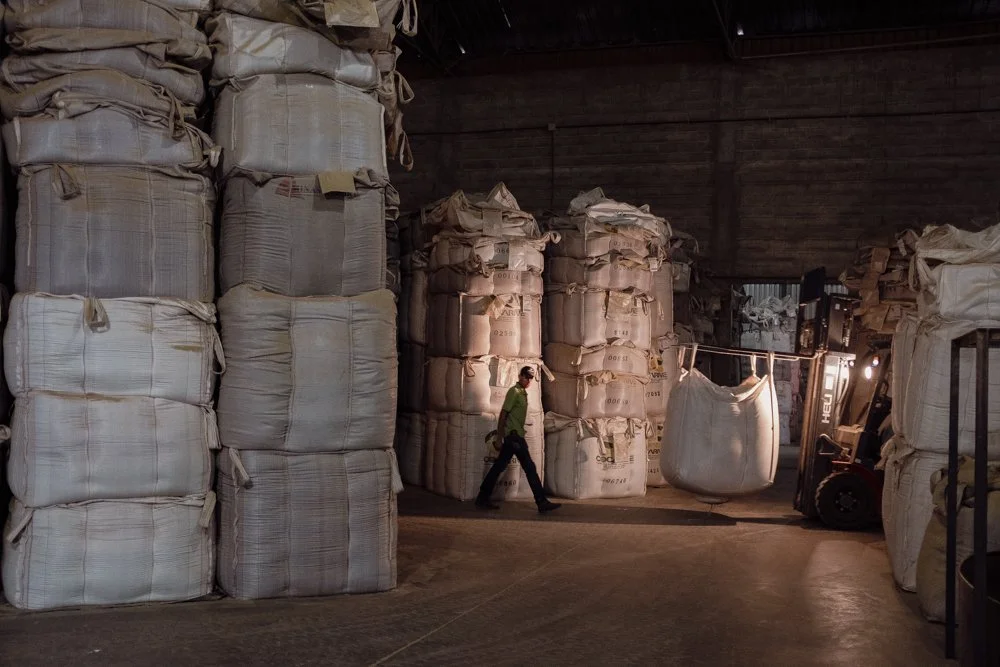A person wearing a green jacket walking through a warehouse with large sacks stacked against the wall, some sacks are open or damaged, and a forklift is visible on the right side.