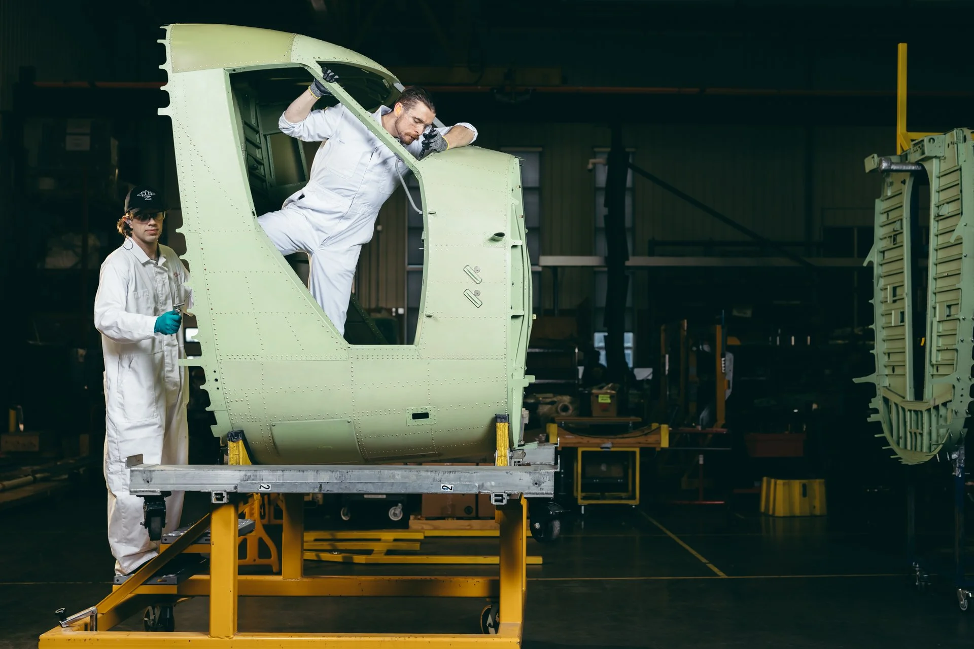 Two workers, one man and one woman, in white coveralls and safety gear, working on assembling an airplane cockpit in a factory. The man is inspecting the interior of the cockpit while the woman holds tools nearby.