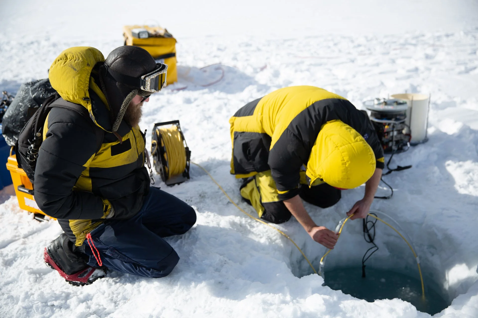 Scientists in yellow and black winter gear working on ice, with equipment and yellow cases nearby, on a snowy landscape.