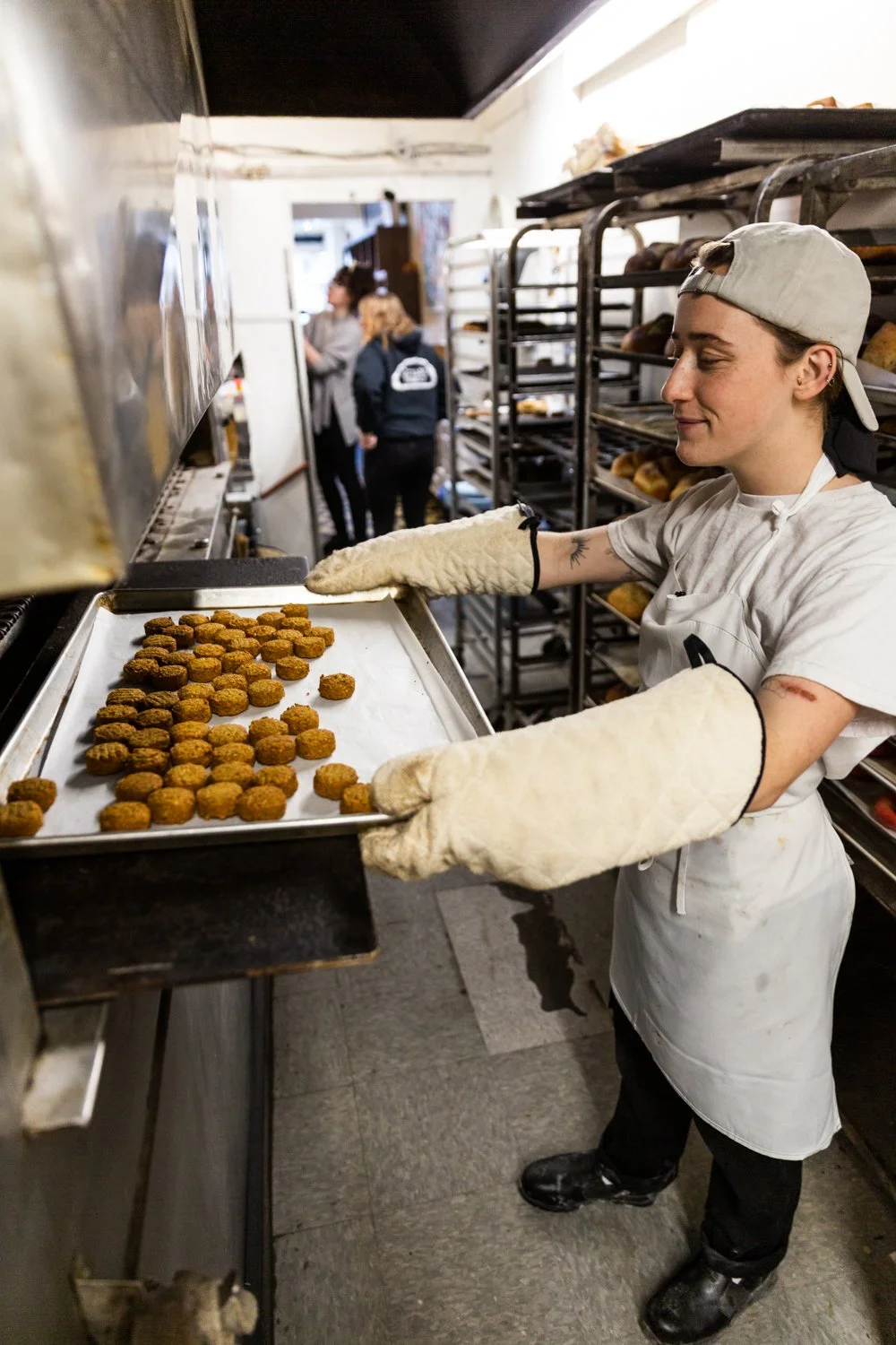 A young woman wearing a gray baseball cap, white chef's coat, and heat-resistant gloves removing a tray of freshly baked chicken nuggets from an oven in a bakery or commercial kitchen. Shelves with baking trays and other workers are visible in the ba