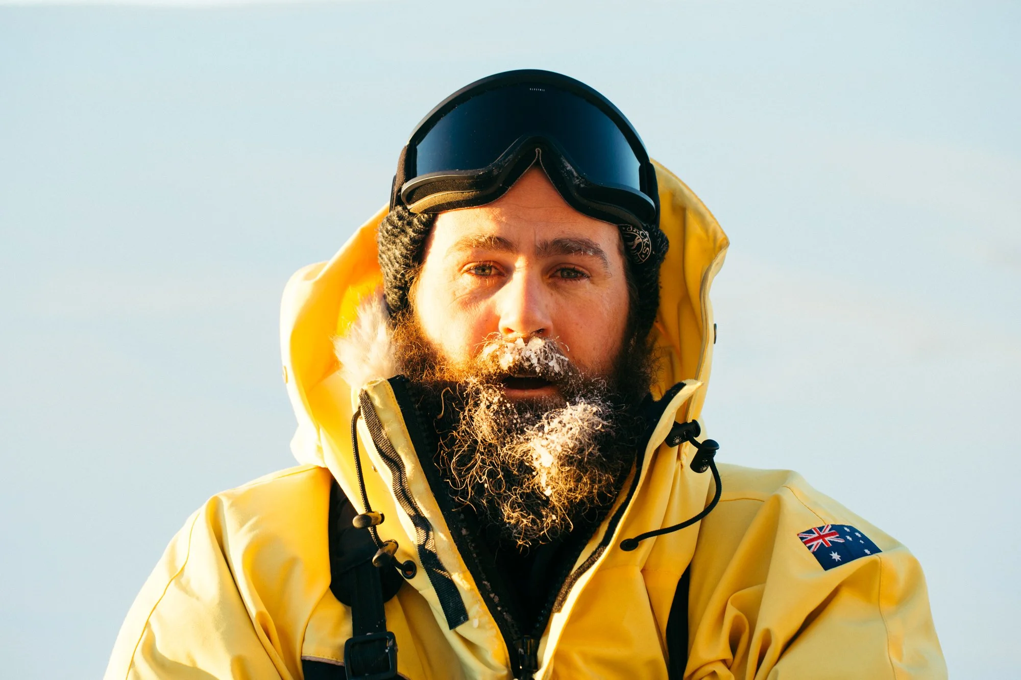 Close-up of a man wearing a yellow winter jacket with an Australian flag patch, black goggles on his forehead, and a gray beanie, outdoors in snowy conditions.