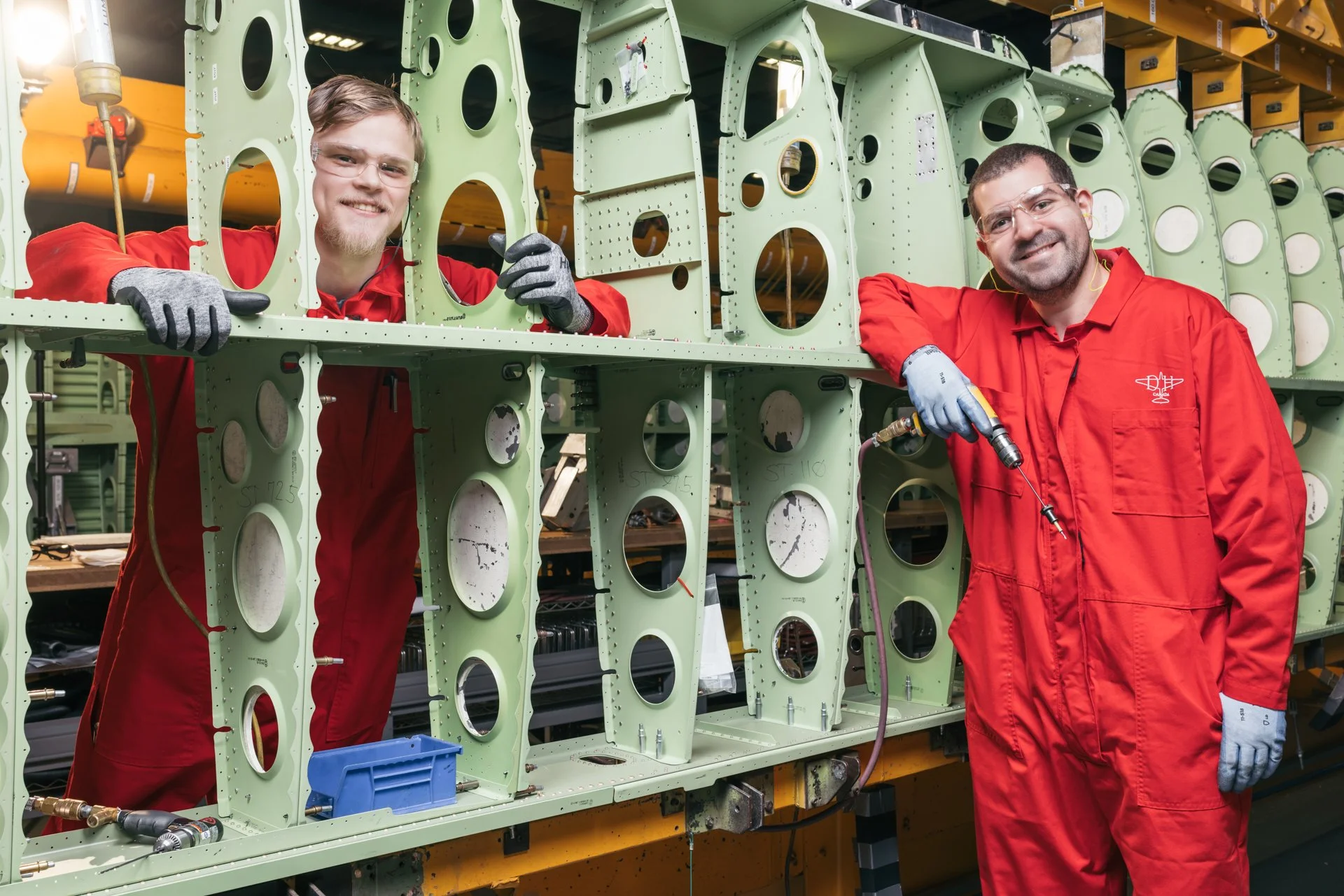 Two workers in red coveralls working on a large piece of aircraft fuselage or metal structure, with one using a tool while smiling at the camera, and the other smiling beside him.