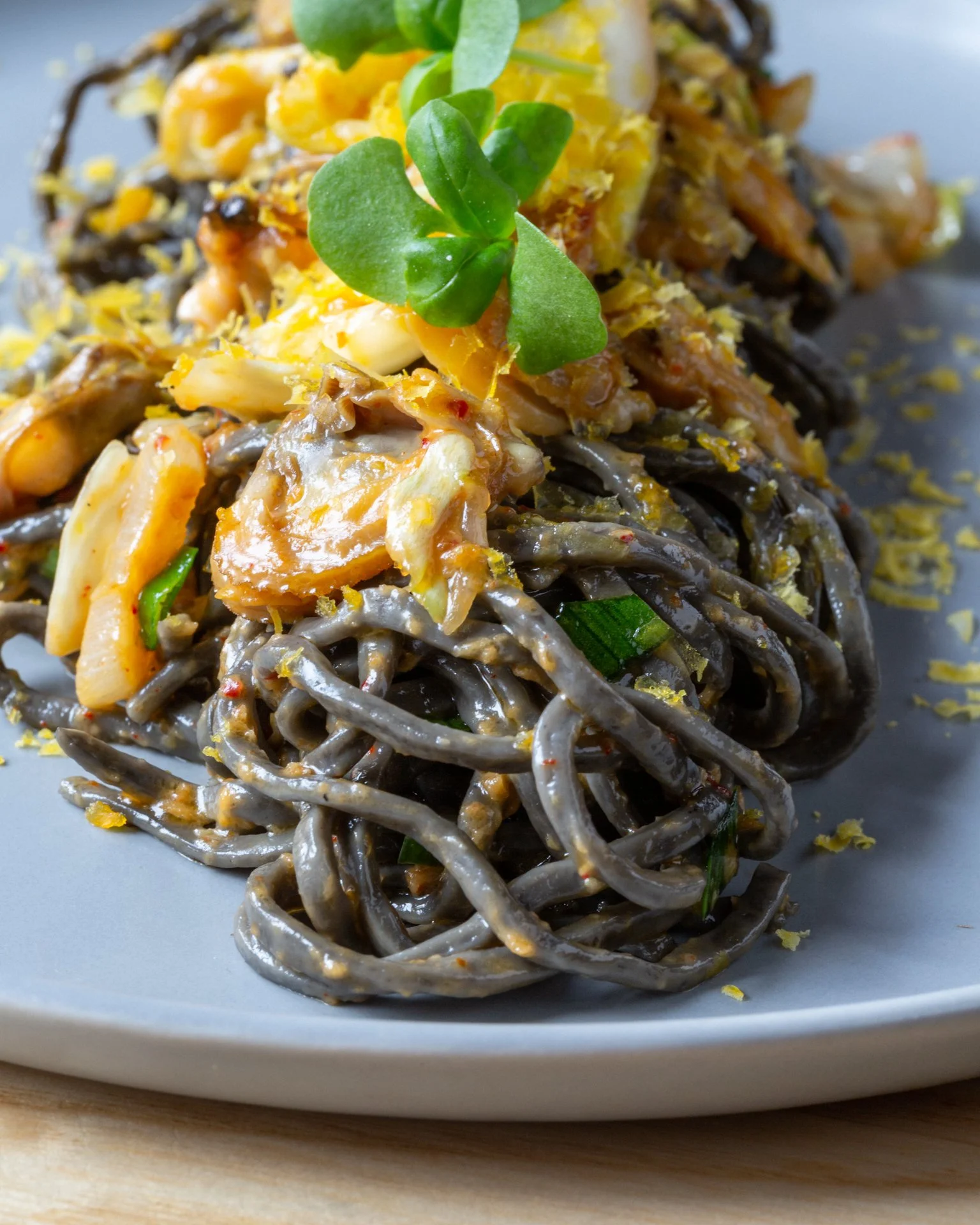 A close-up of black noodles topped with shredded cheese, pieces of cooked chicken, chopped green onions, and garnished with fresh microgreens, served on a white plate.