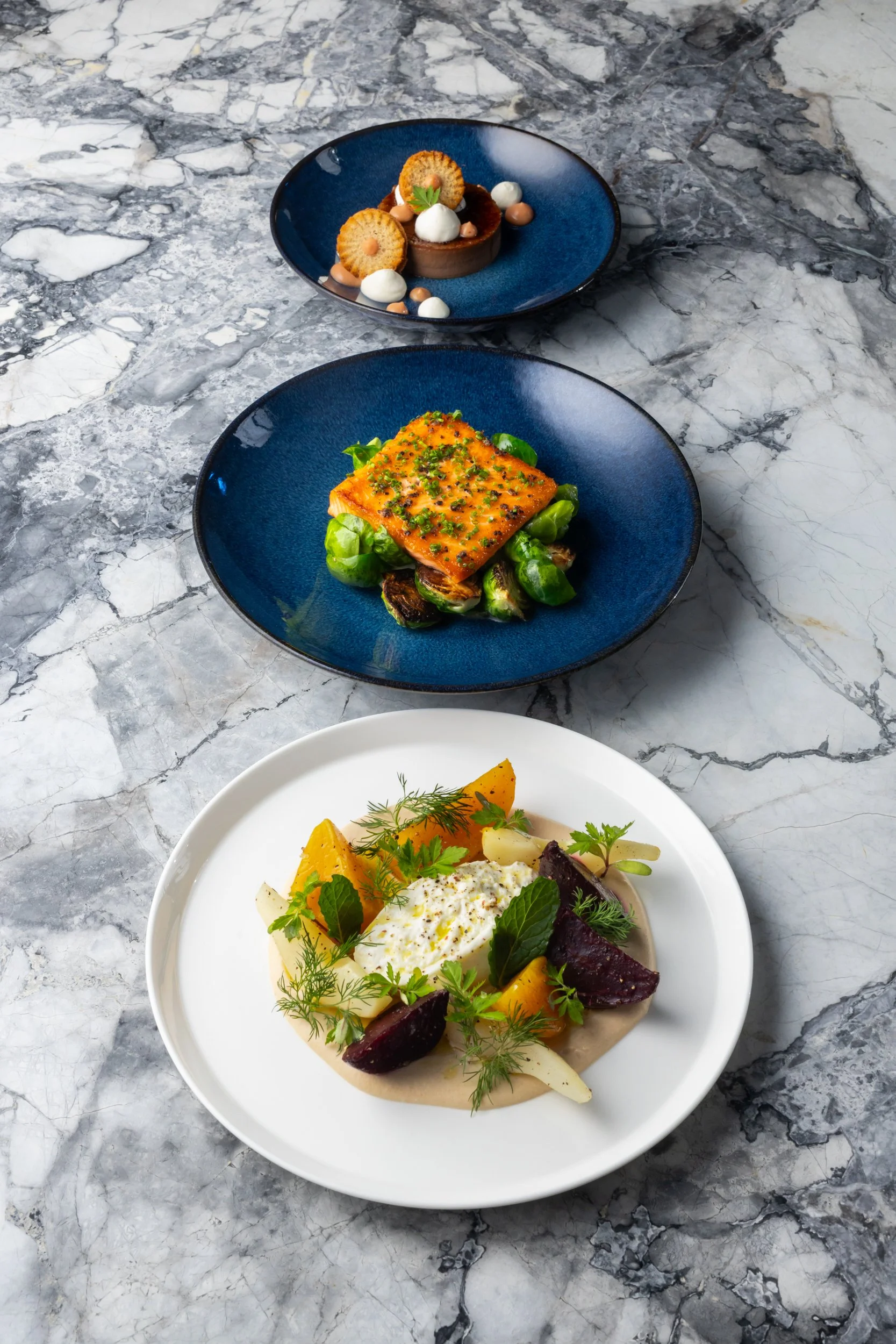 Three elegant plates of gourmet food on a marble table. The top plate features a chocolate tart decorated with small cookies, dollops of cream, and a sprig of parsley. The middle plate contains a piece of glazed salmon on a bed of Brussels sprouts, g