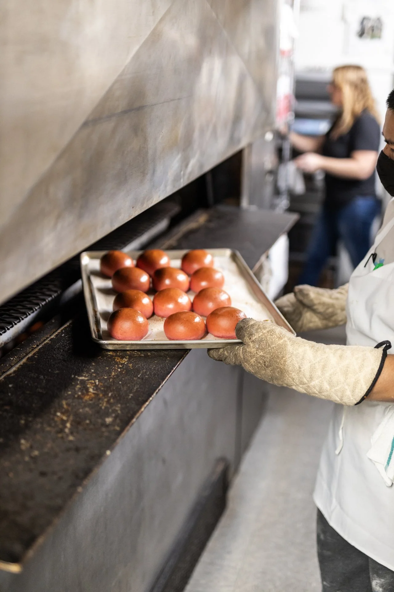 A person wearing gloves and a white apron is holding a tray of baked red round pastries or buns in a commercial kitchen. In the background, other staff members are working.