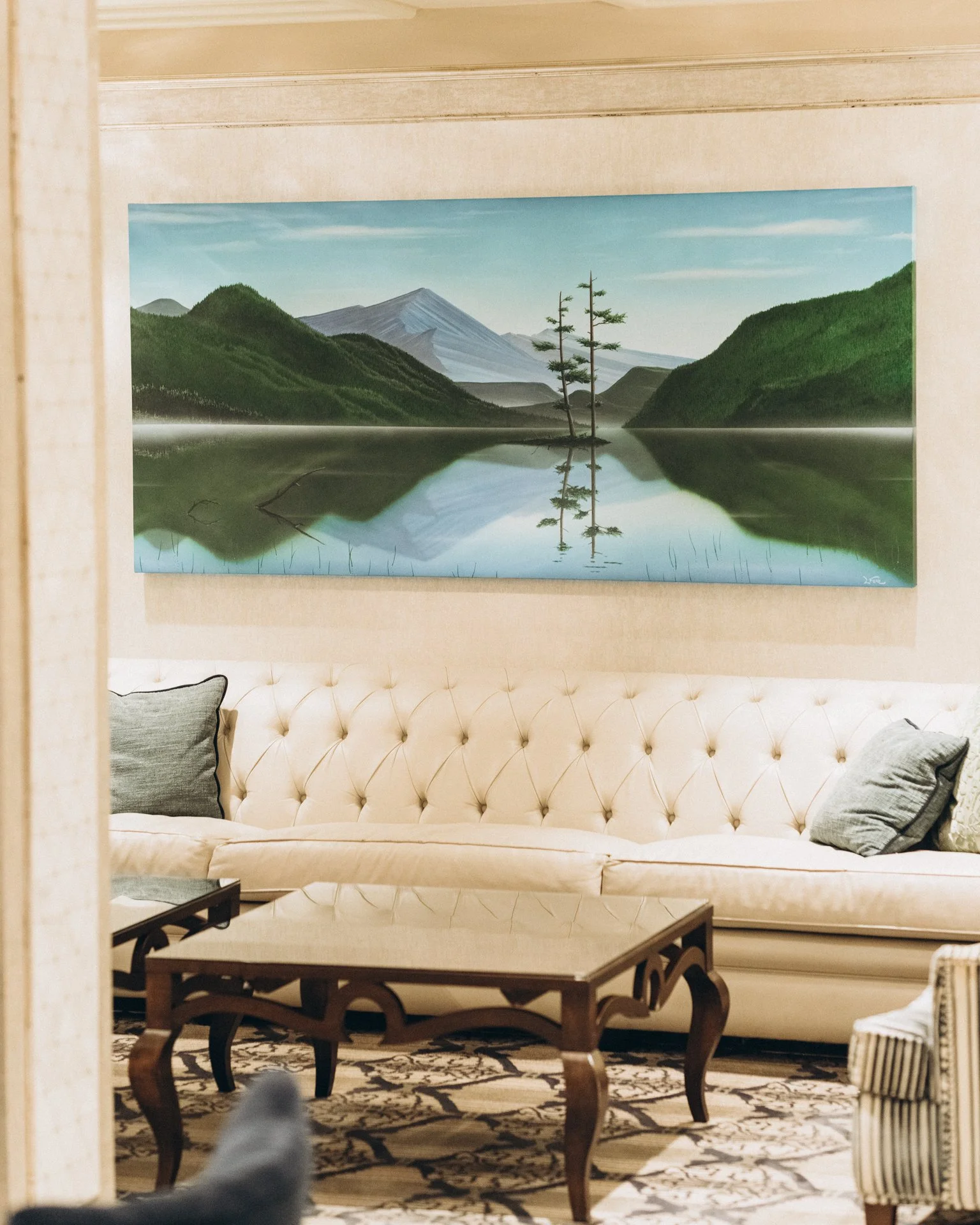 A landscape painting depicts mountains, trees, and a lake reflecting the scenery, hanging on a beige wall above a cream tufted velvet sofa with gray pillows in a living room. There is a wooden coffee table with curved legs in front of the sofa.