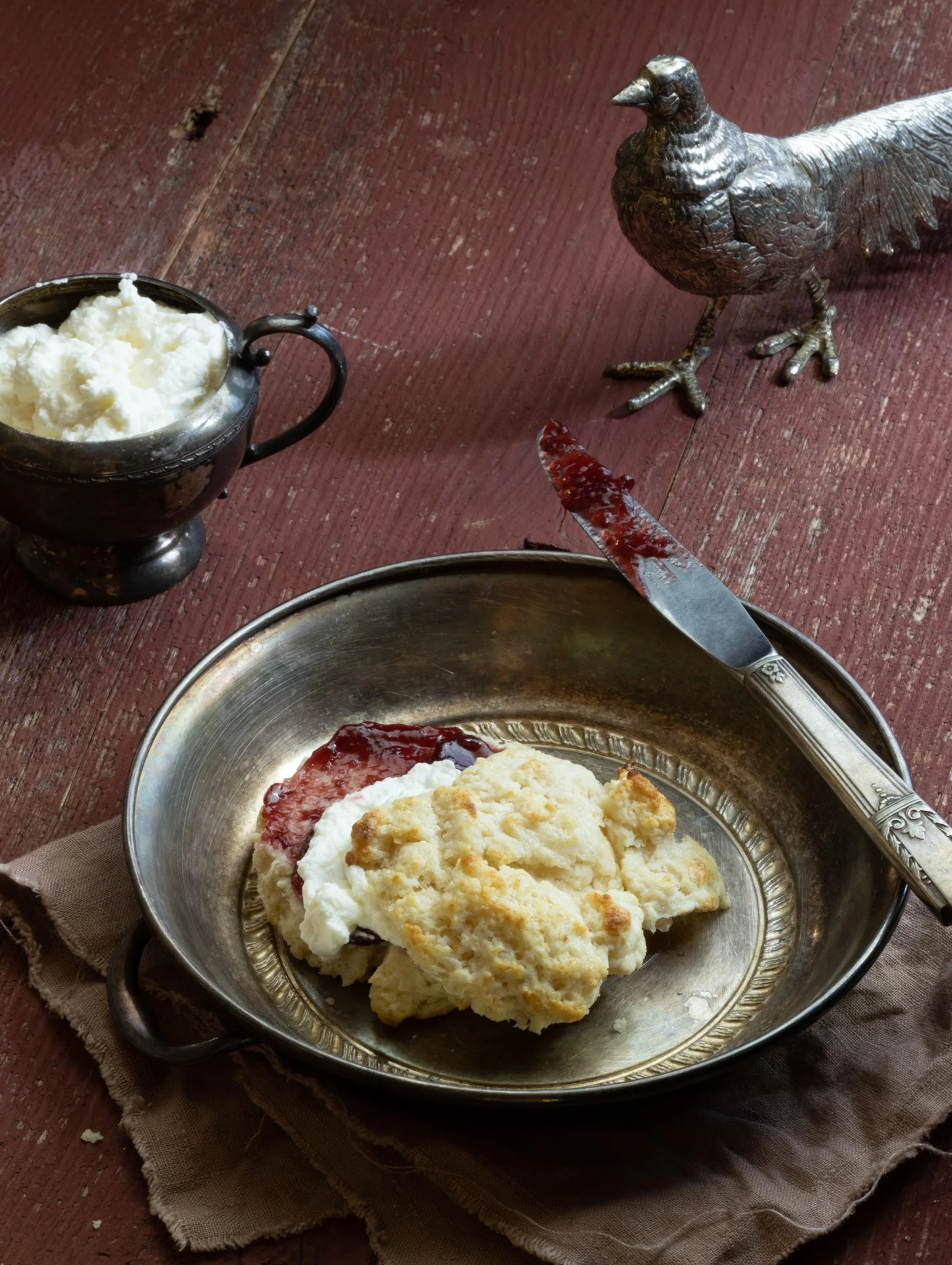A metal plate with mashed potatoes and cherry or strawberry jam, on a beige cloth napkin. There is a butter knife with jam on it resting on the plate. In the background, there is a small metal container filled with whipped butter, a decorative metal 