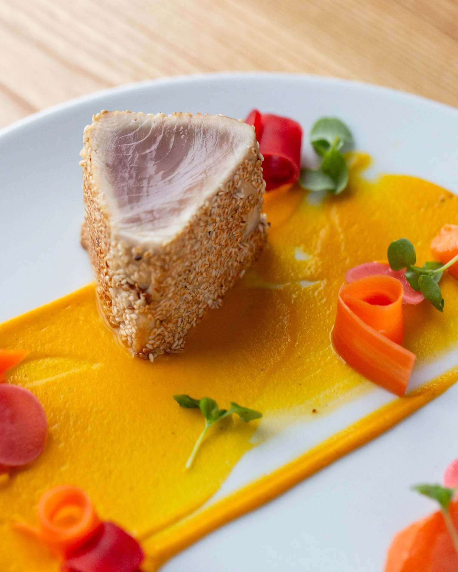 Close-up of a plated dish featuring a piece of sesame-crusted tuna on a bed of bright orange puree, garnished with colorful vegetables, including curly slices of carrot, round slices of radish, and small greens, on a white plate.