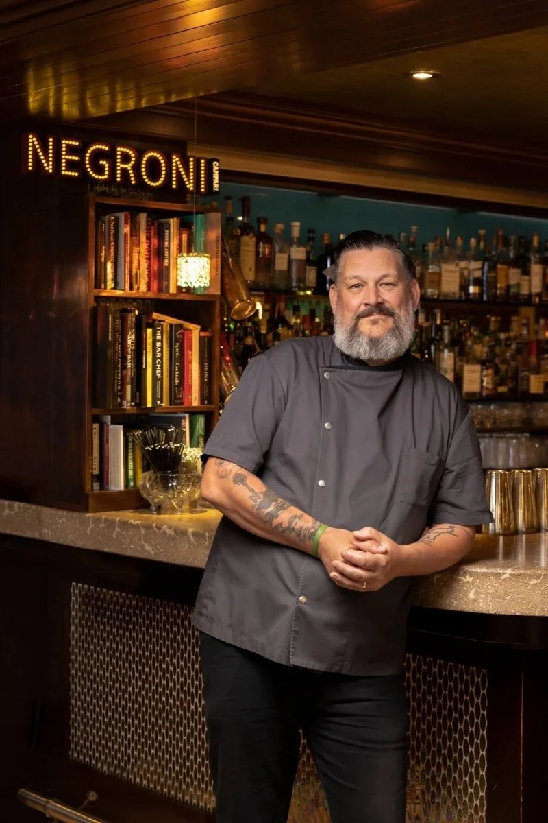 A chef with a beard and tattoos standing behind a bar with liquor bottles, books, and a glow sign that reads 'NEGRONI' in a restaurant or bar setting.