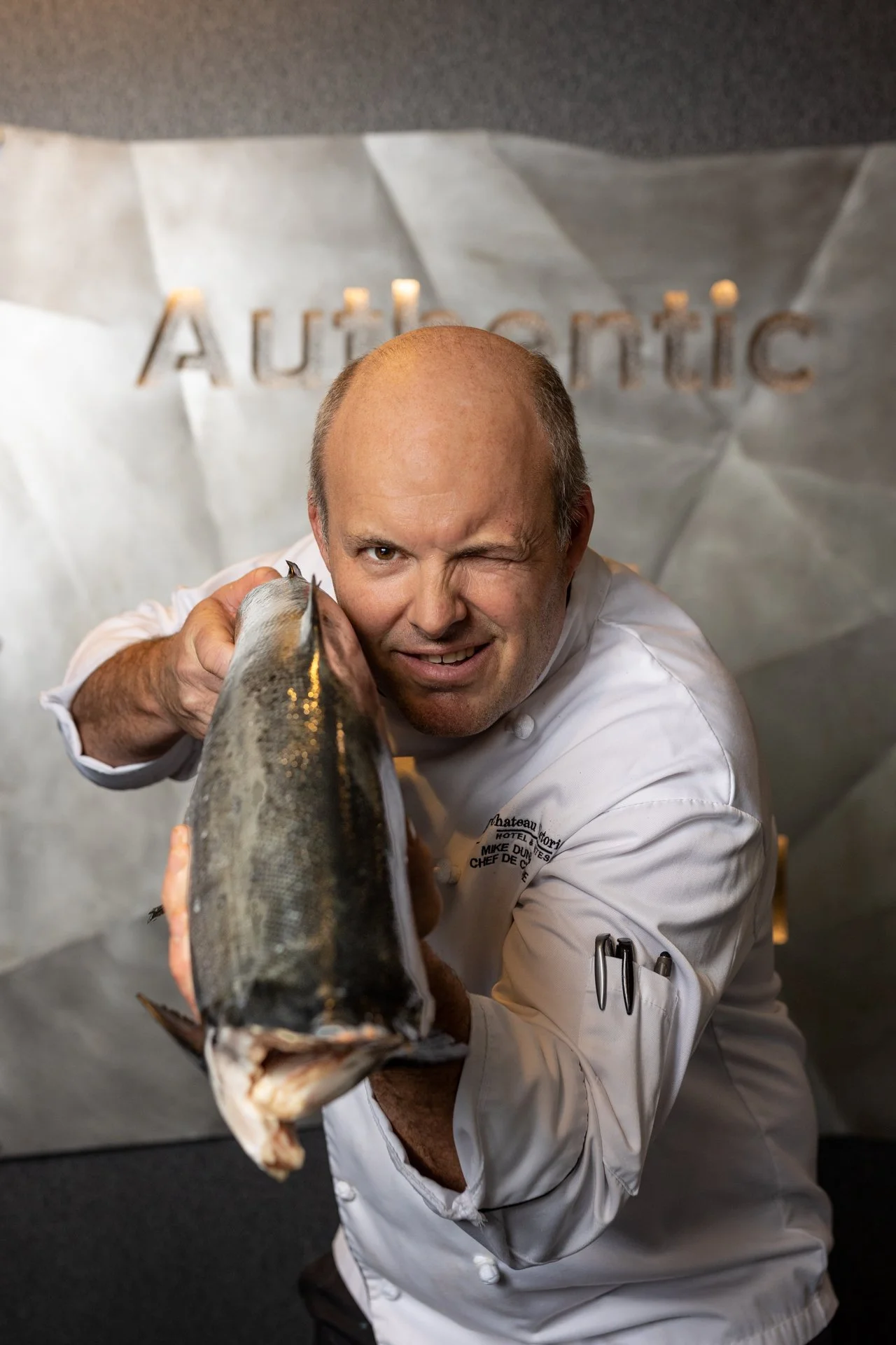 A chef in a white uniform holding a large fish towards the camera in a professional kitchen with a metallic background that has the word 'Authentic' on it.
